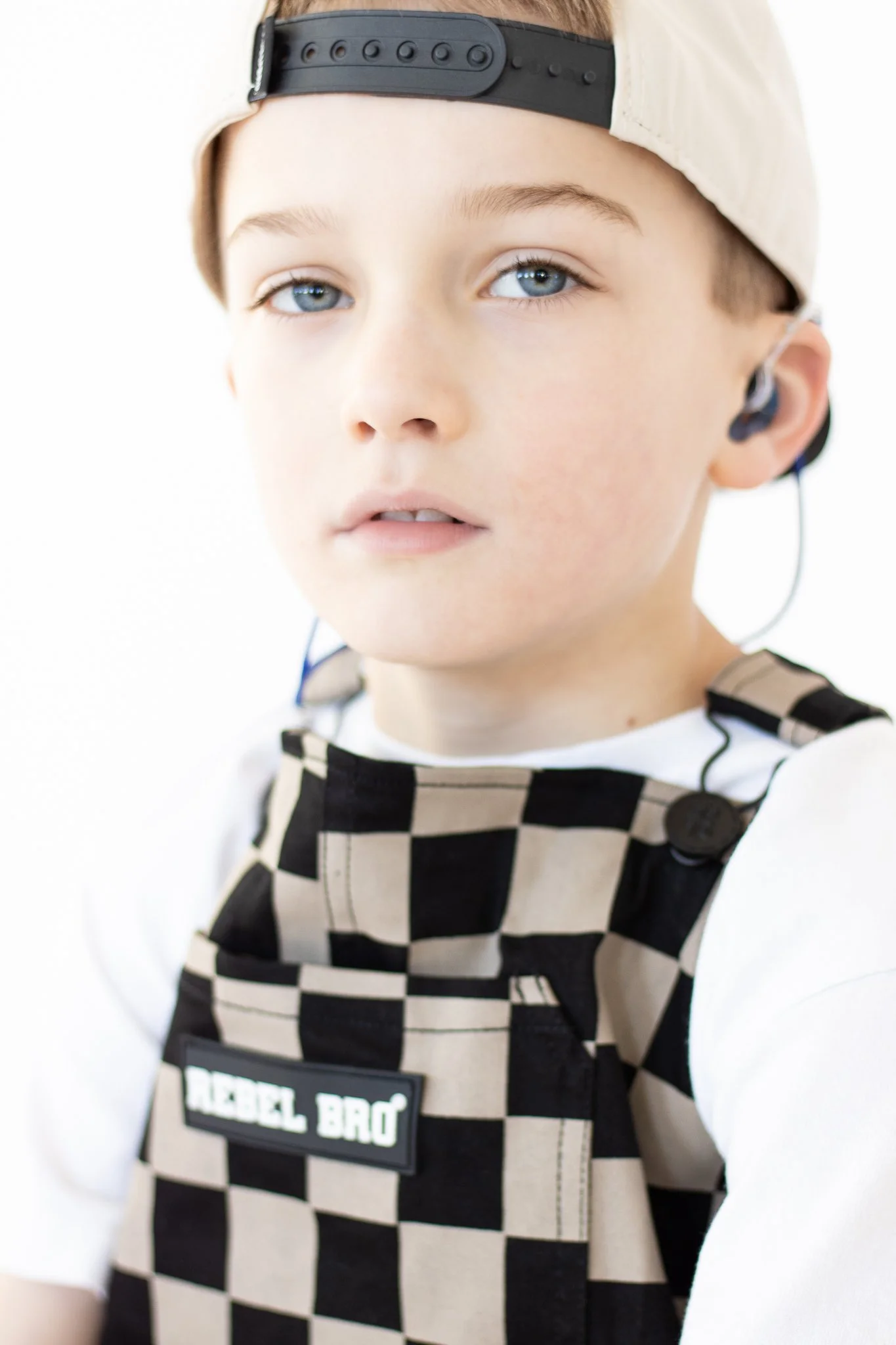 A young boy wearing a beige cap backward, a white shirt, and a black and beige checkered overall labeled "REBEL BRO". He has blue eyes and is wearing an earpiece in his right ear, looking slightly to the side.