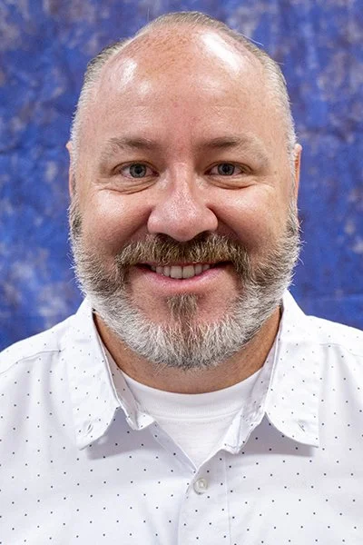 A smiling man with glasses and a beard wearing a green shirt that says "Receive love" in a room with wooden cabinets.