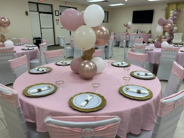 Round banquet tables with pink tablecloths and white covers on chairs, decorated with pink, white, and rose gold balloons in centerpieces for a celebration or party.