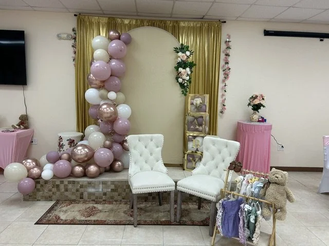 Decorative stage setup with a balloon arrangement of pink, white, and metallic balloons, a backdrop with golden curtains, floral accents, two white tufted chairs, a small rack with clothes and a teddy bear, and a pink table with flowers.