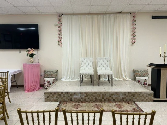 Indoor stage with two white tufted chairs, floral decorations, pink drape, small platform, and floral arrangements, set for a ceremony or event.