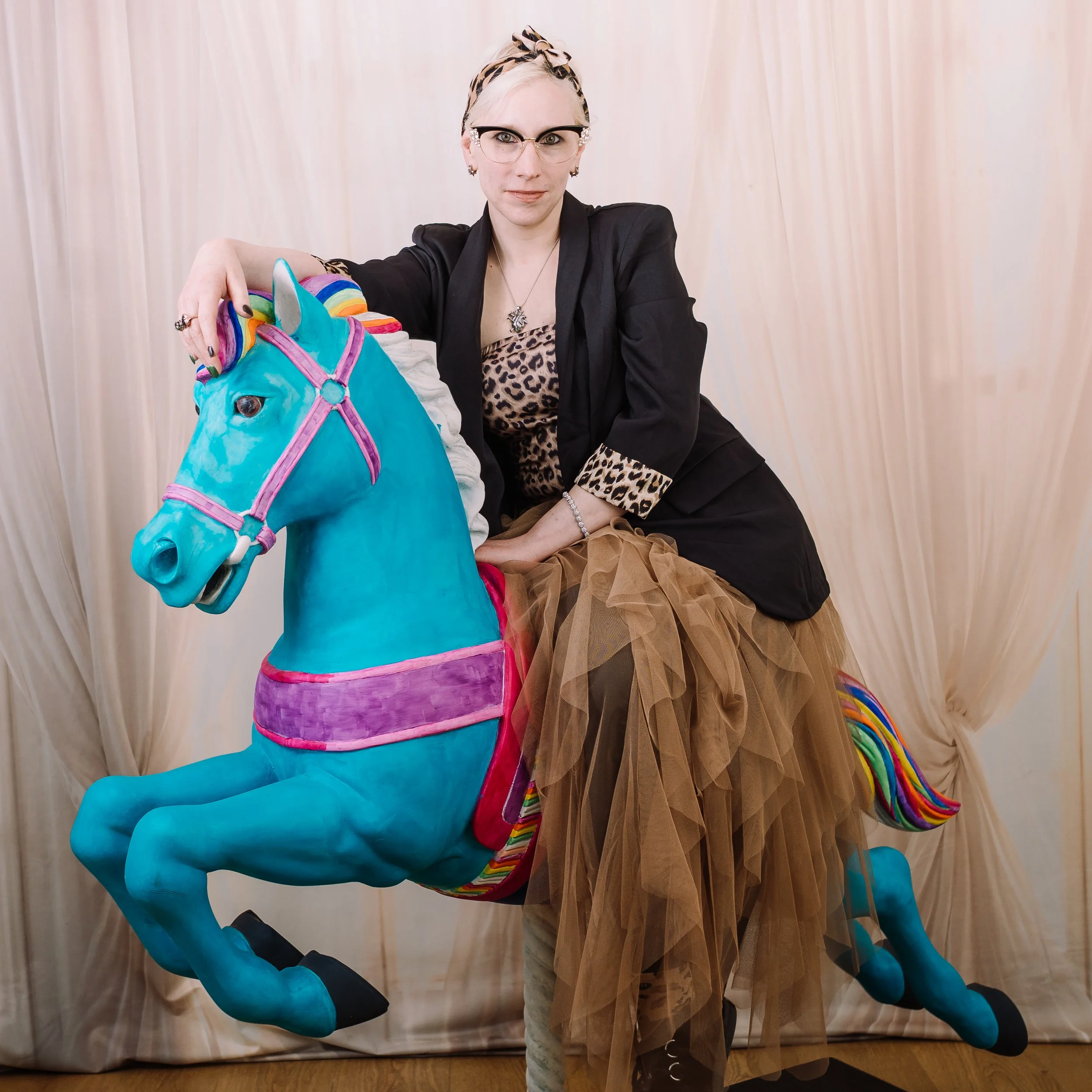 A woman wearing glasses, leopard print top, black blazer, and a brown tulle skirt sits on a blue carousel horse with pink, purple, and rainbow accents, posed in front of a beige curtain backdrop.