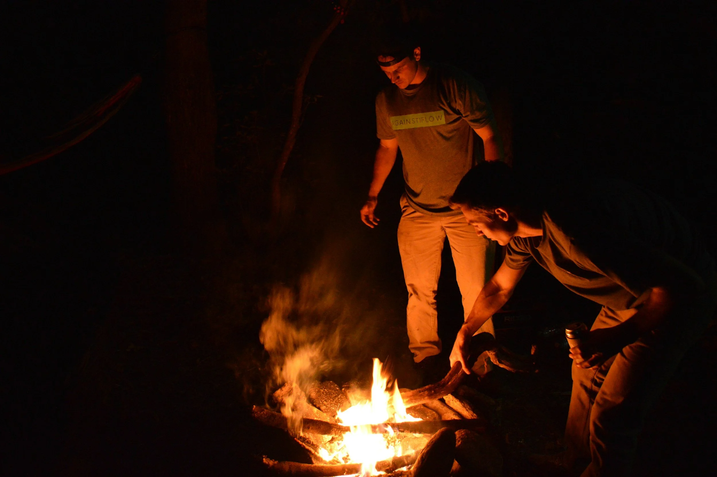 Two young men are tending to a campfire at night outdoors. One is bending down, holding a log, while the other stands nearby watching. The scene is dimly lit by the fire.