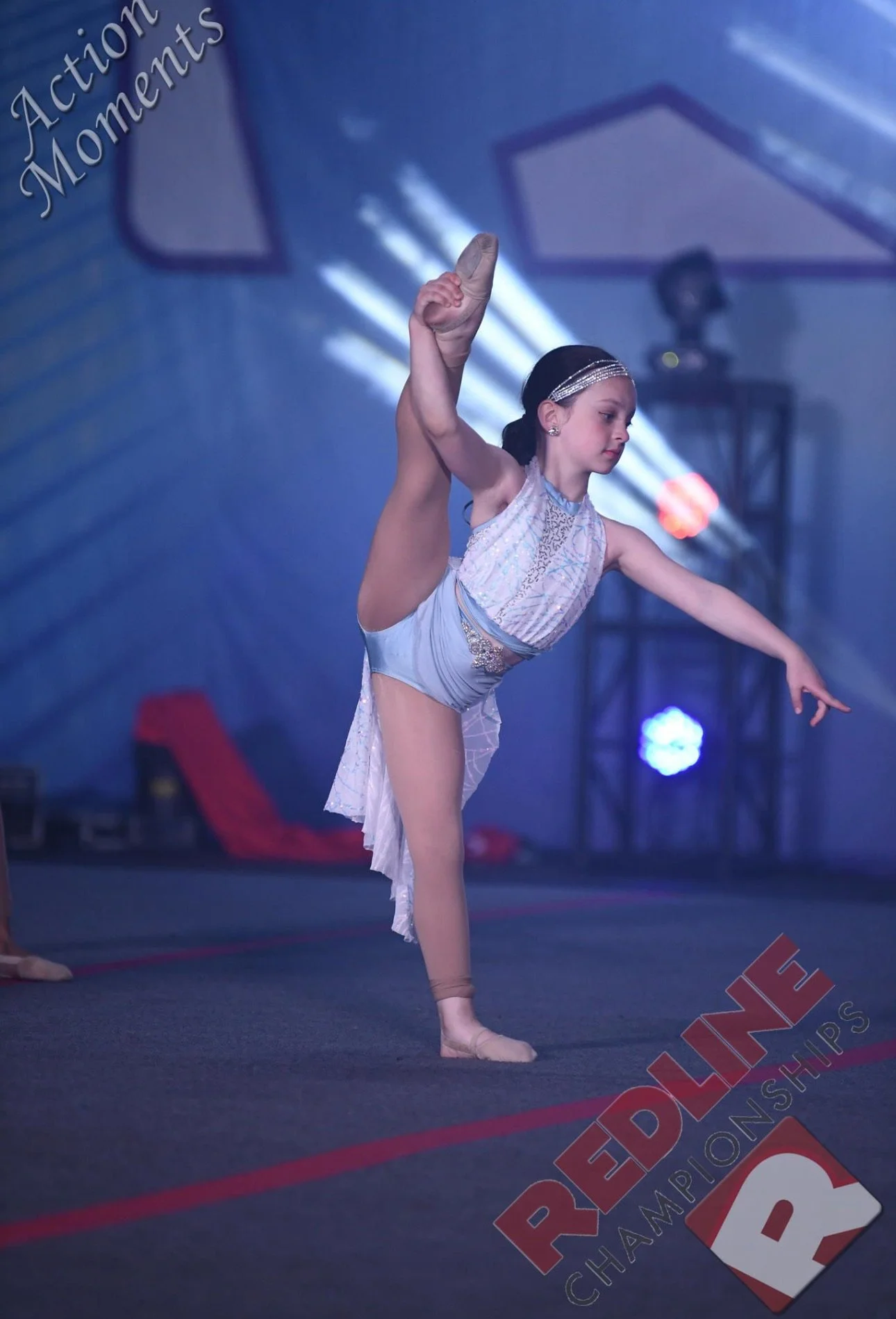 Young girl in a light-colored dance costume performs an advanced dance move on stage at a competition, with the REDLINE Championships logo visible on the floor.
