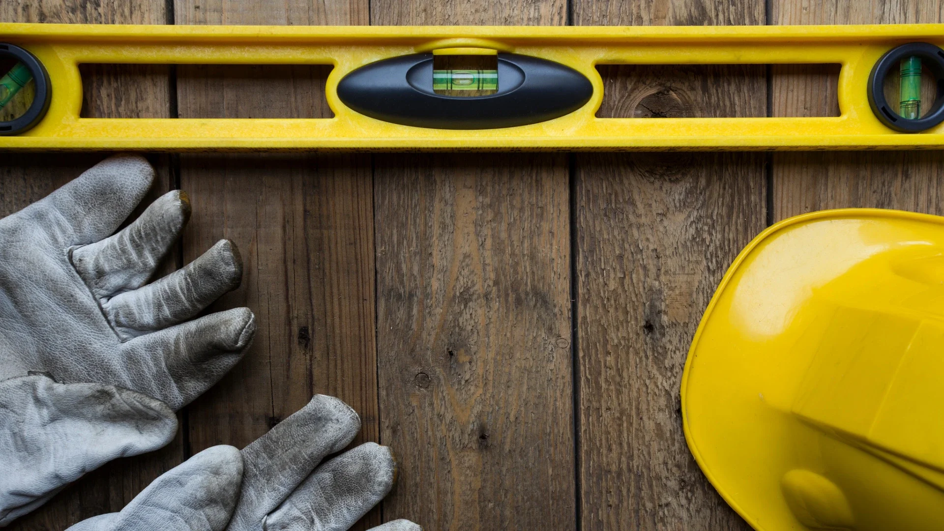 Yellow spirit level, white work gloves, and yellow construction helmet arranged on a wooden surface.