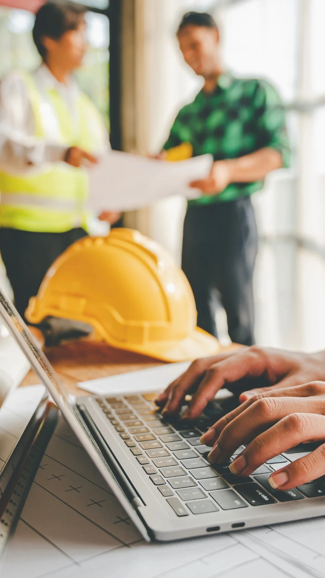 Close-up of hands typing on a laptop keyboard at a work site with a yellow safety helmet and construction plans nearby, while two people in construction vests and casual clothing discuss in the background.