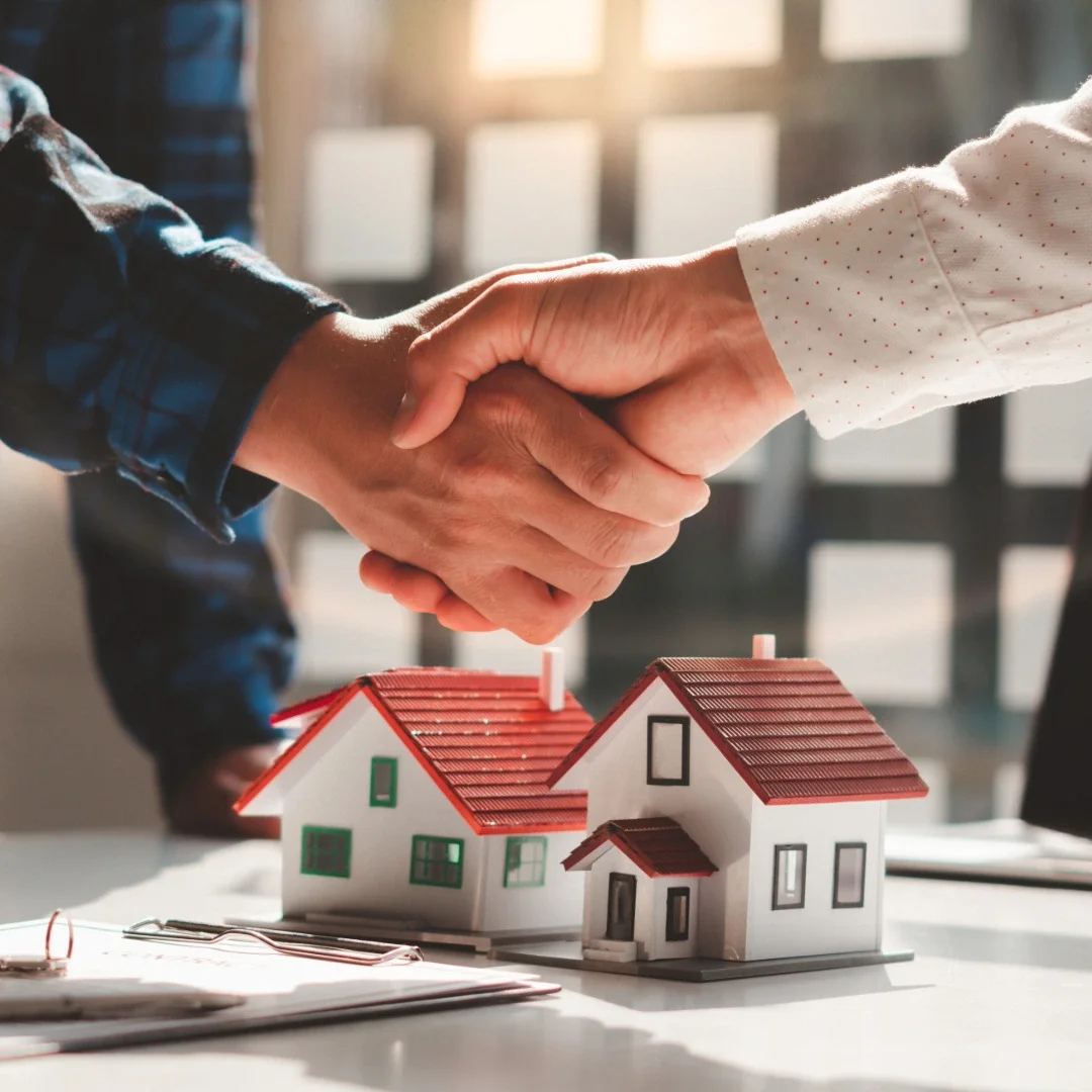 Two people shaking hands over a table with miniature houses, symbolizing a real estate agreement or property deal.