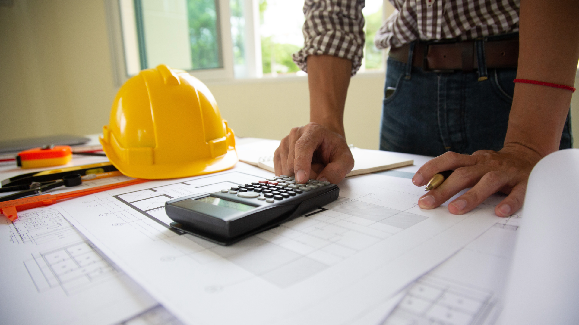 Person working on architectural plans with a yellow safety helmet, calculator, and drafting tools on a table in a well-lit room.