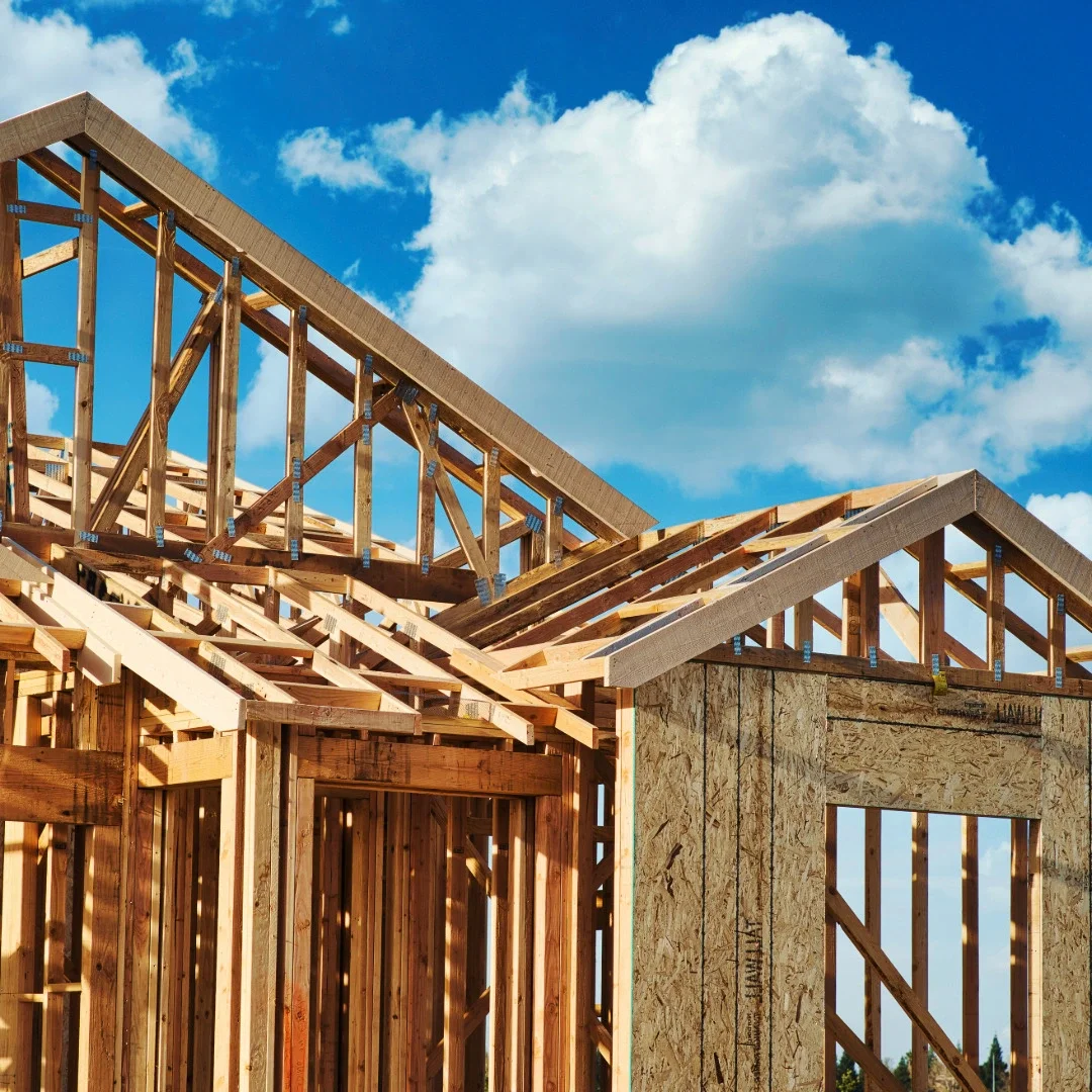 Wooden house under construction with visible framing, roof trusses, and a blue sky with clouds