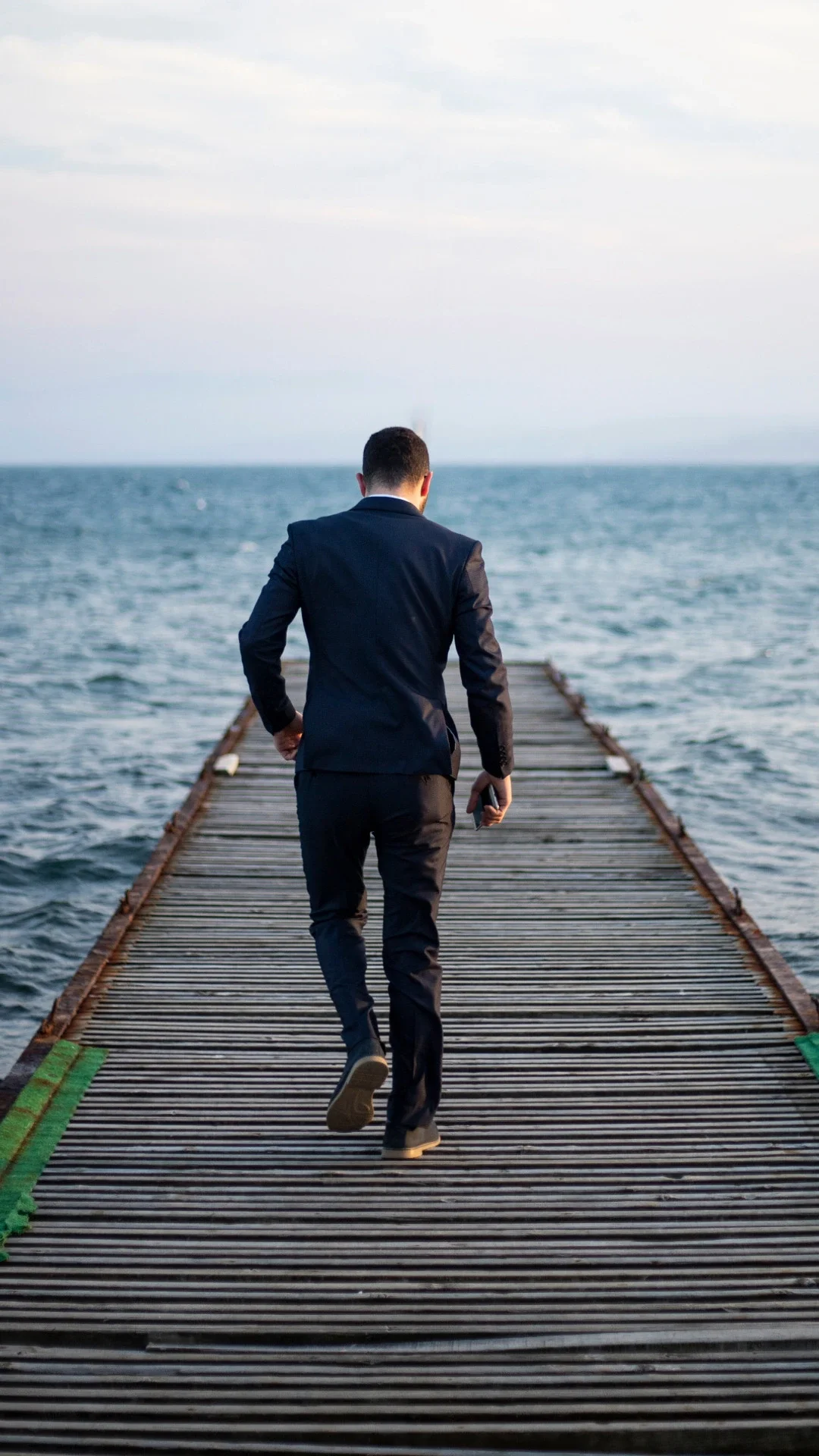 A man in a black suit walking on a wooden dock over water, holding a phone in his right hand.