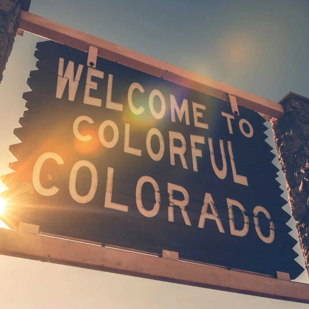 A sign reading 'Welcome to Colorful Colorado' with the sun shining behind it, mounted on a wooden frame with stone pillars.