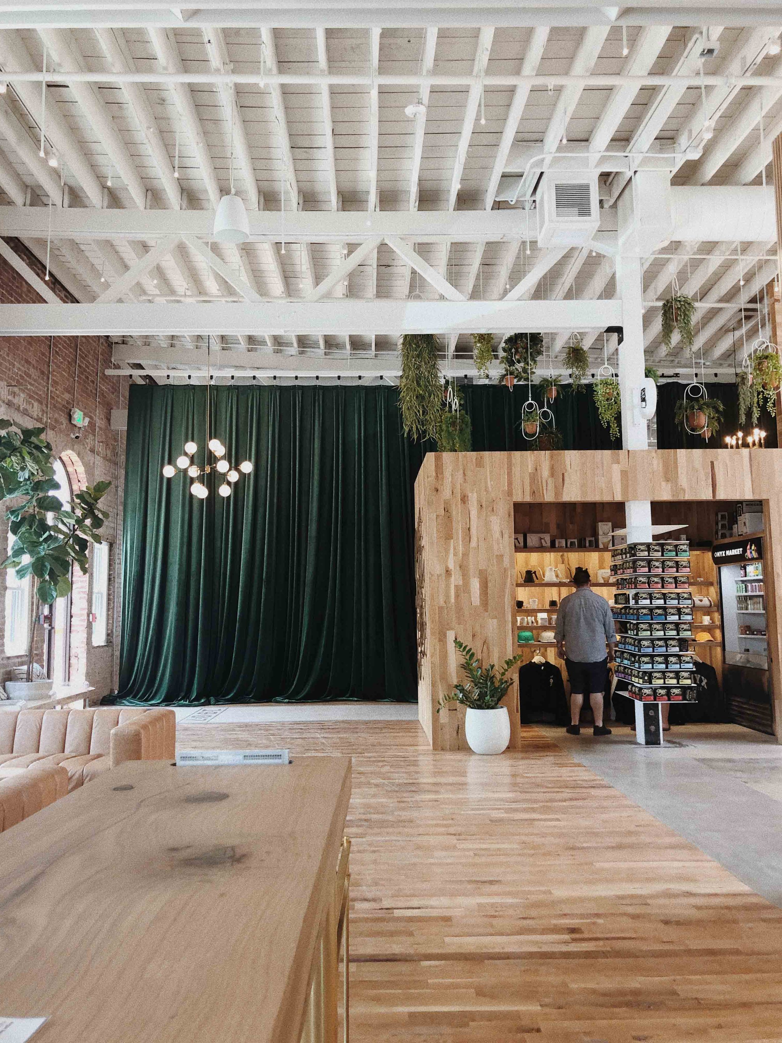 Interior de un espacio moderno con paredes de ladrillo, cortinas verdes y plantas colgantes, con una zona de bar y una estructura de madera.