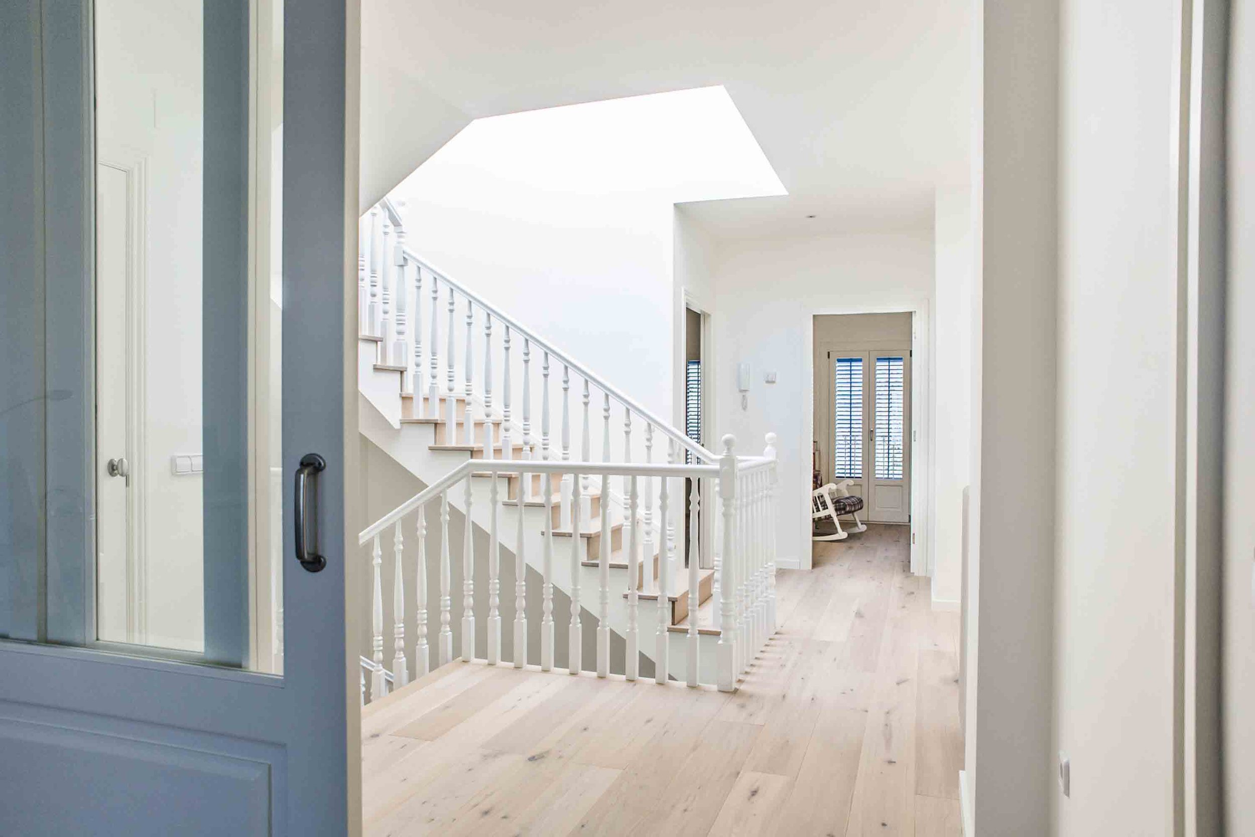 Entrada a una casa con escalera de madera y barandales blancos, paredes blancas y piso de madera natural, con puertas de vidrio con contraventanas de viento hacia el exterior.