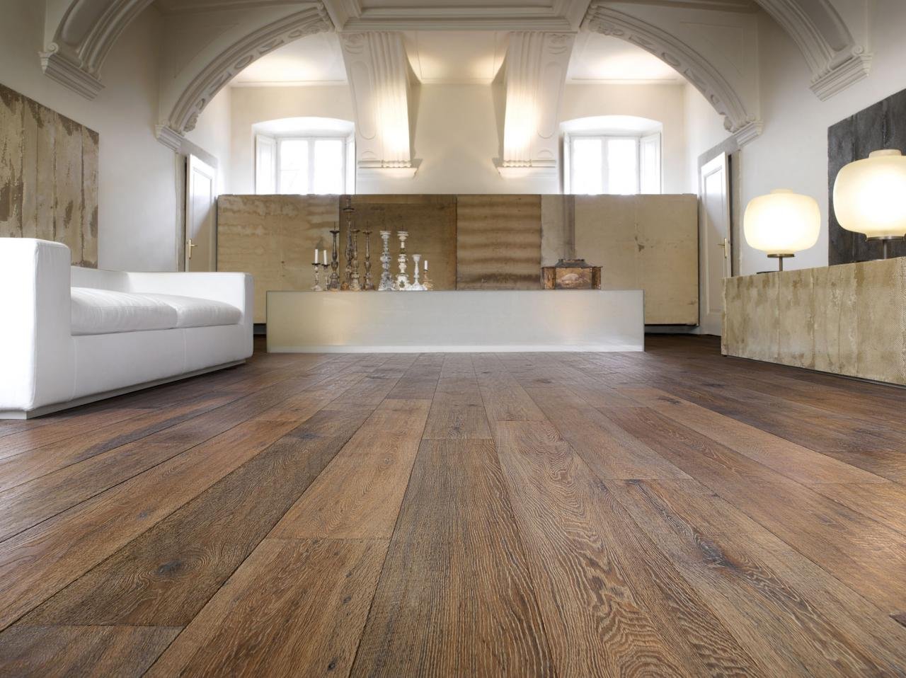 Sala con suelo de madera, sillón blanco, lámparas de mesa, y decoración minimalista en un espacio amplio con ventanas y techos con molduras.