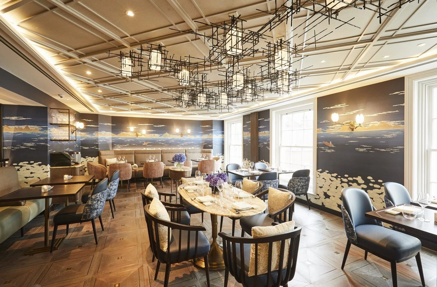 Un restaurante elegante con paredes decoradas con un mural que simula un paisaje acuático y muebles de diferentes estilos, incluyendo mesas de mármol y sillas tapizadas en tonos neutros.