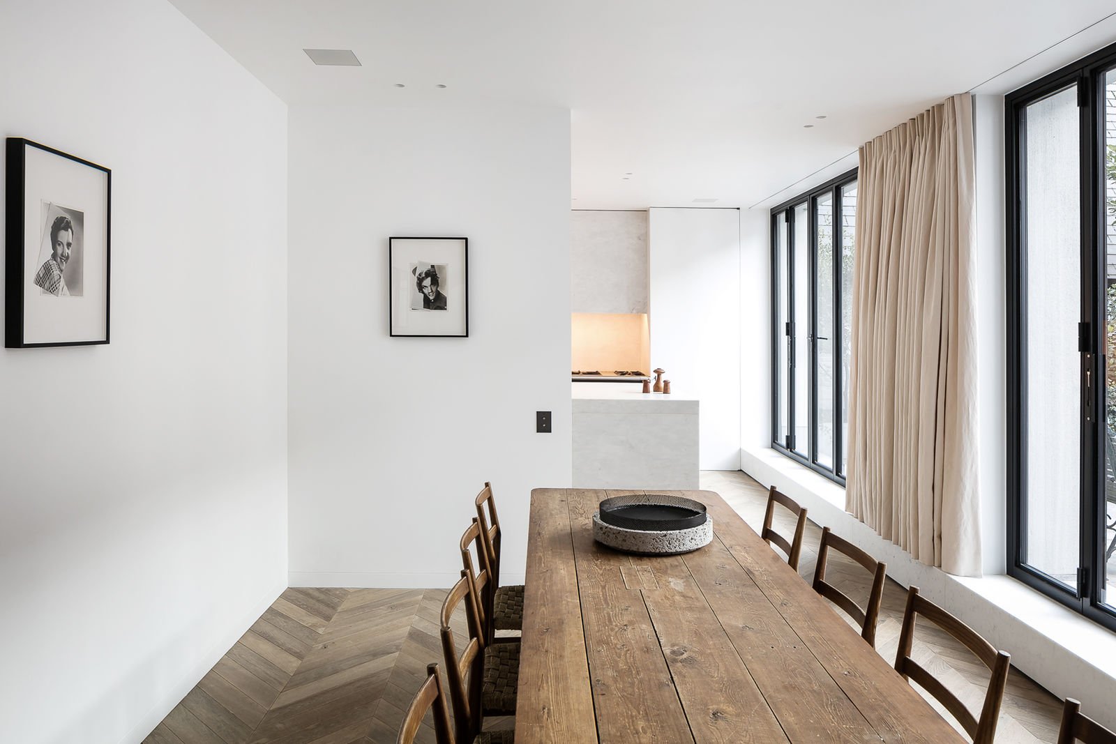 Sala de comedor con mesa de madera y sillas, grandes ventanas con cortinas beige, paredes blancas, fotos enmarcadas en la pared, cocina en el fondo, luz natural