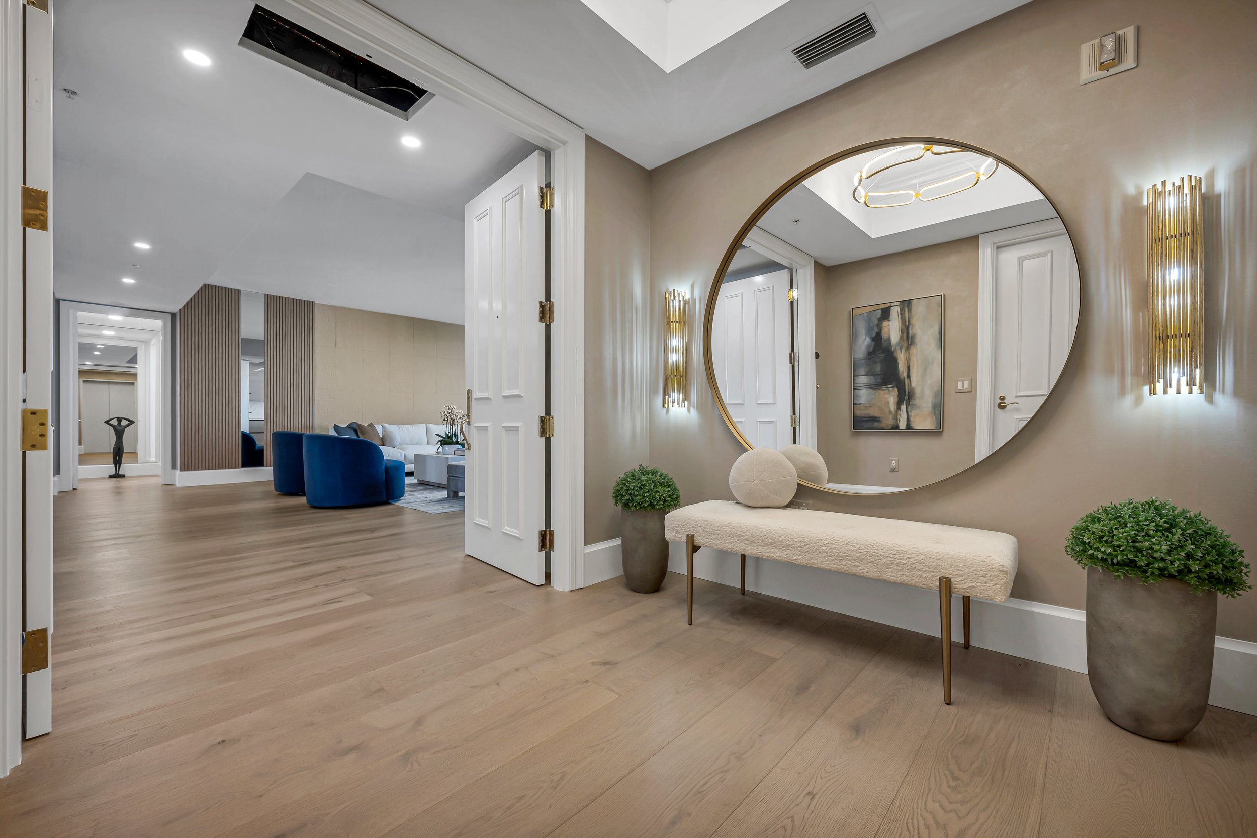 Vestíbulo decorado con un gran espejo circular, dos macetas con plantas verdes, una banca con cojines y paredes con arte y lámparas de pared