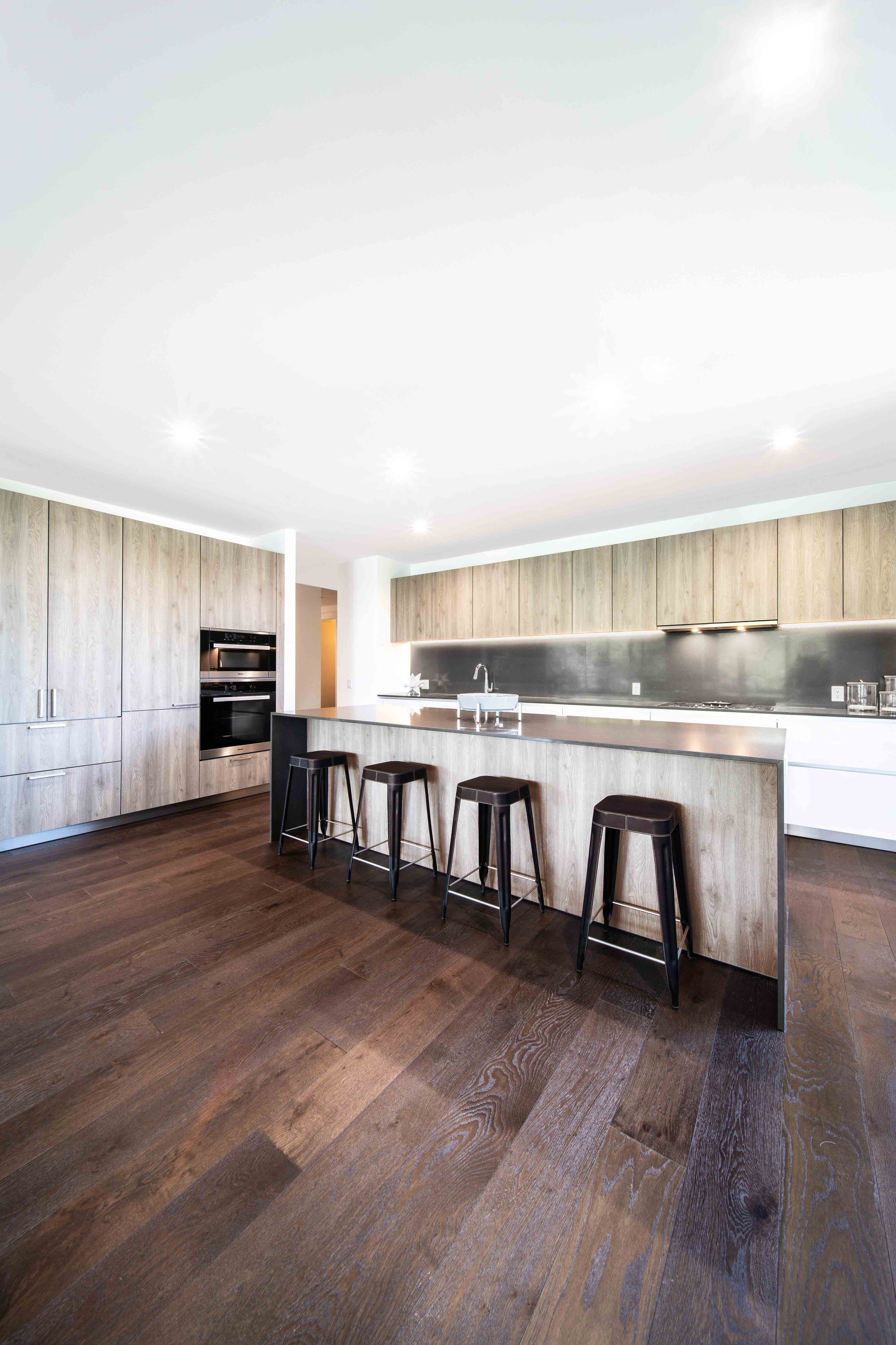 Cocina moderna con gabinetes de madera y encimera de piedra, barra para comer con cuatro sillas negras, piso de madera oscuro, paredes blancas y techos con luces empotradas.
