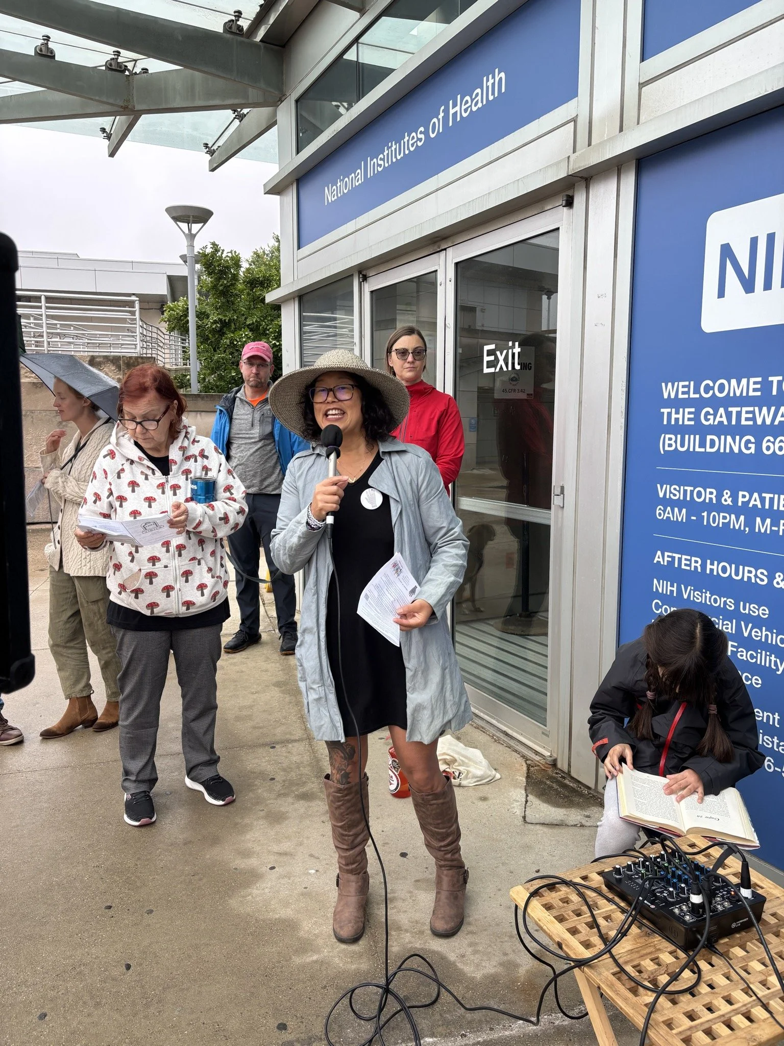 Smiling speaker in hat and blue jacket holds microphone in front of NIH entrance doors
