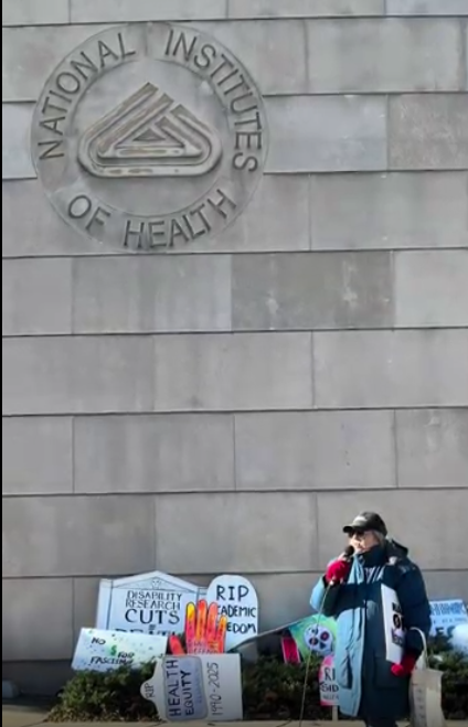 A person speaking at an NIH vigil, with the NIH logo visible on the wall above thema