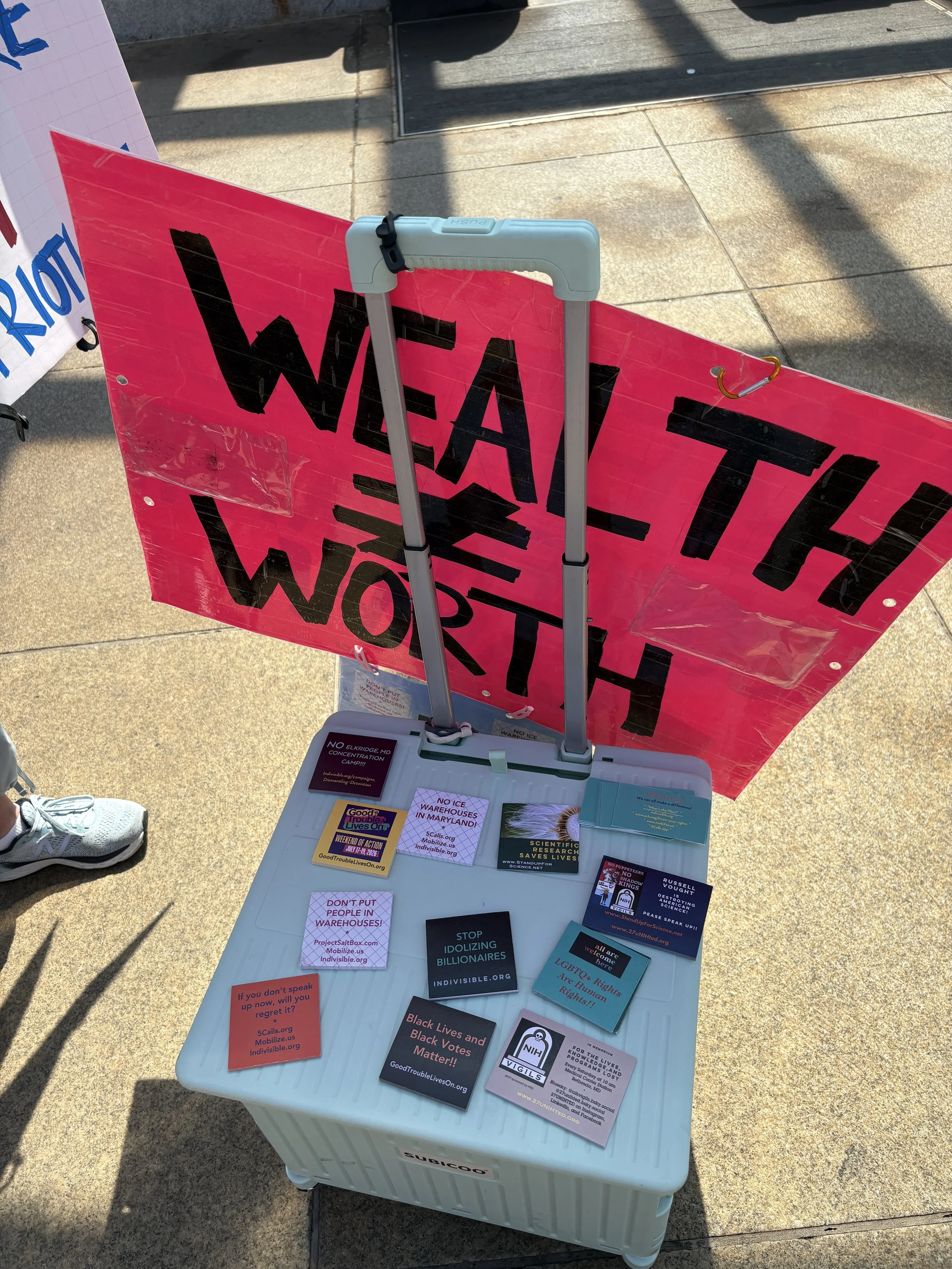 A rolling supply bin displays advocacy handouts beneath a sign reading “Wealth ≠ Worth.”