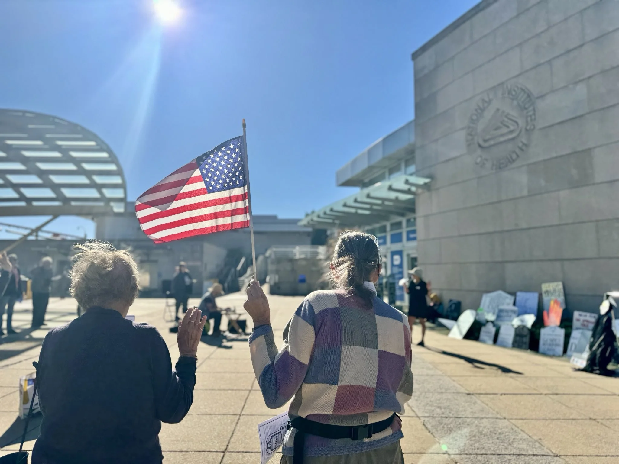 A person, seen from behind, holds their right hand up to recite the Oath of Office while the person next to her waves an American flag. Both are standing outside NIH at a vigil.