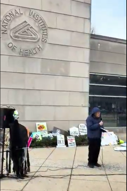 A person speaks into a microphone at an NIH vigil