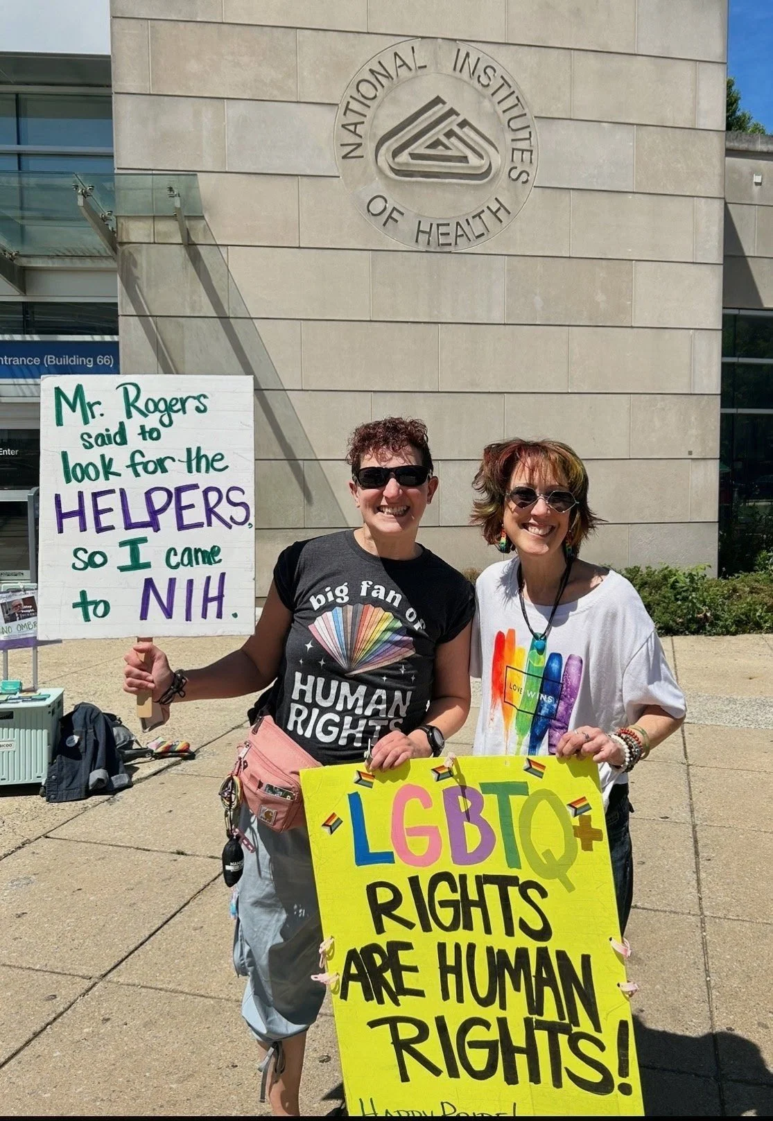 Two NIH Vigil attendees hold signs supporting helpers and LGBTQ+ rights outside the NIH entrance.