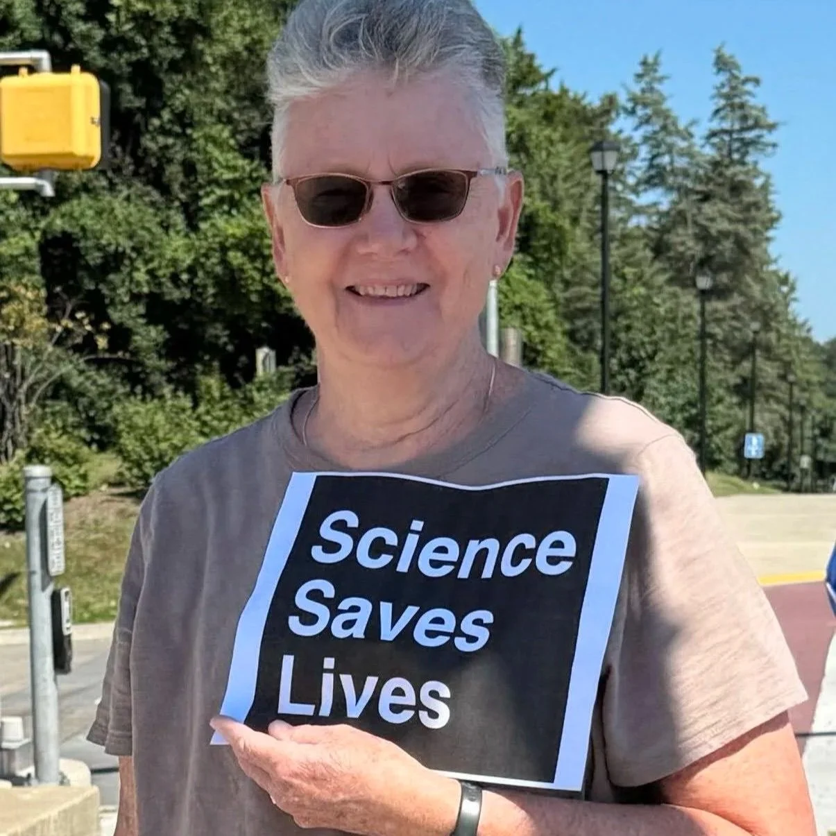 A woman with sunglasses and a beige T-shirt, holding a small sign that says Science Saves Lives.