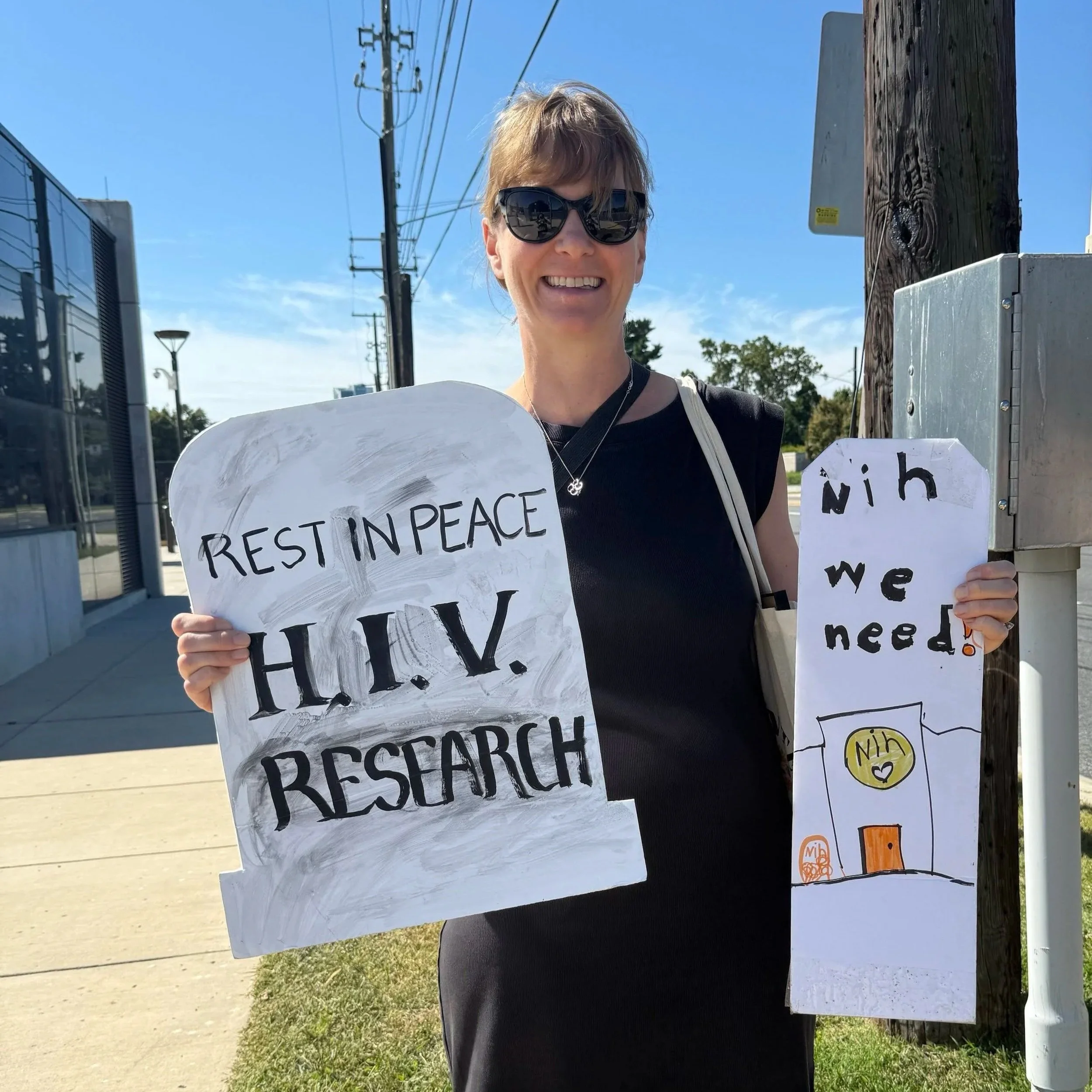 A woman in a black dress holding two signs. One is shaped like a tombstone and says Rest in Peace HIV research and the other says NIH we need.