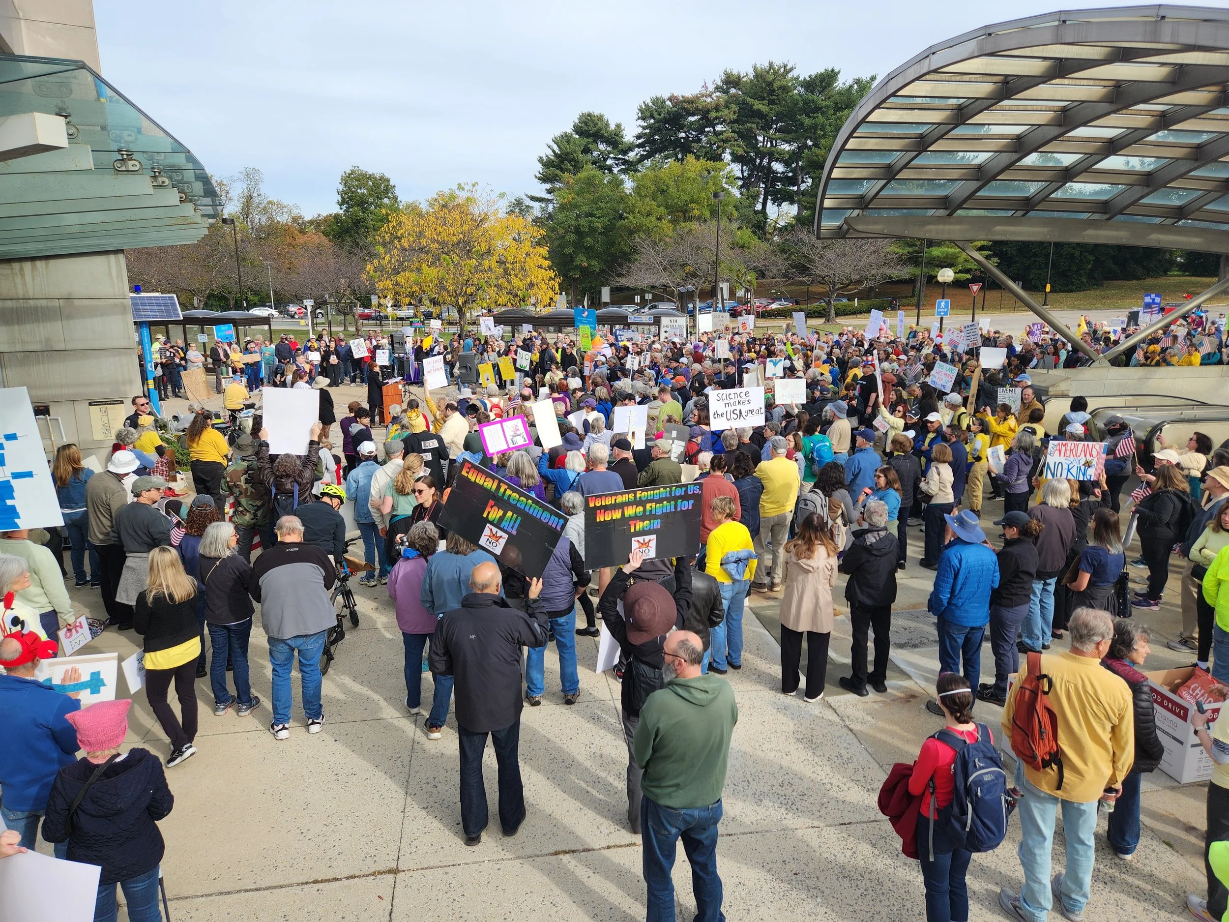 A large group of people standing near the entrance to the NIH metro station, many holding signs to show their support for science research and funding