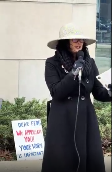 A protest sign behind a vigil speaker, saying "Dear feds, we appreciate you! Your work is important."