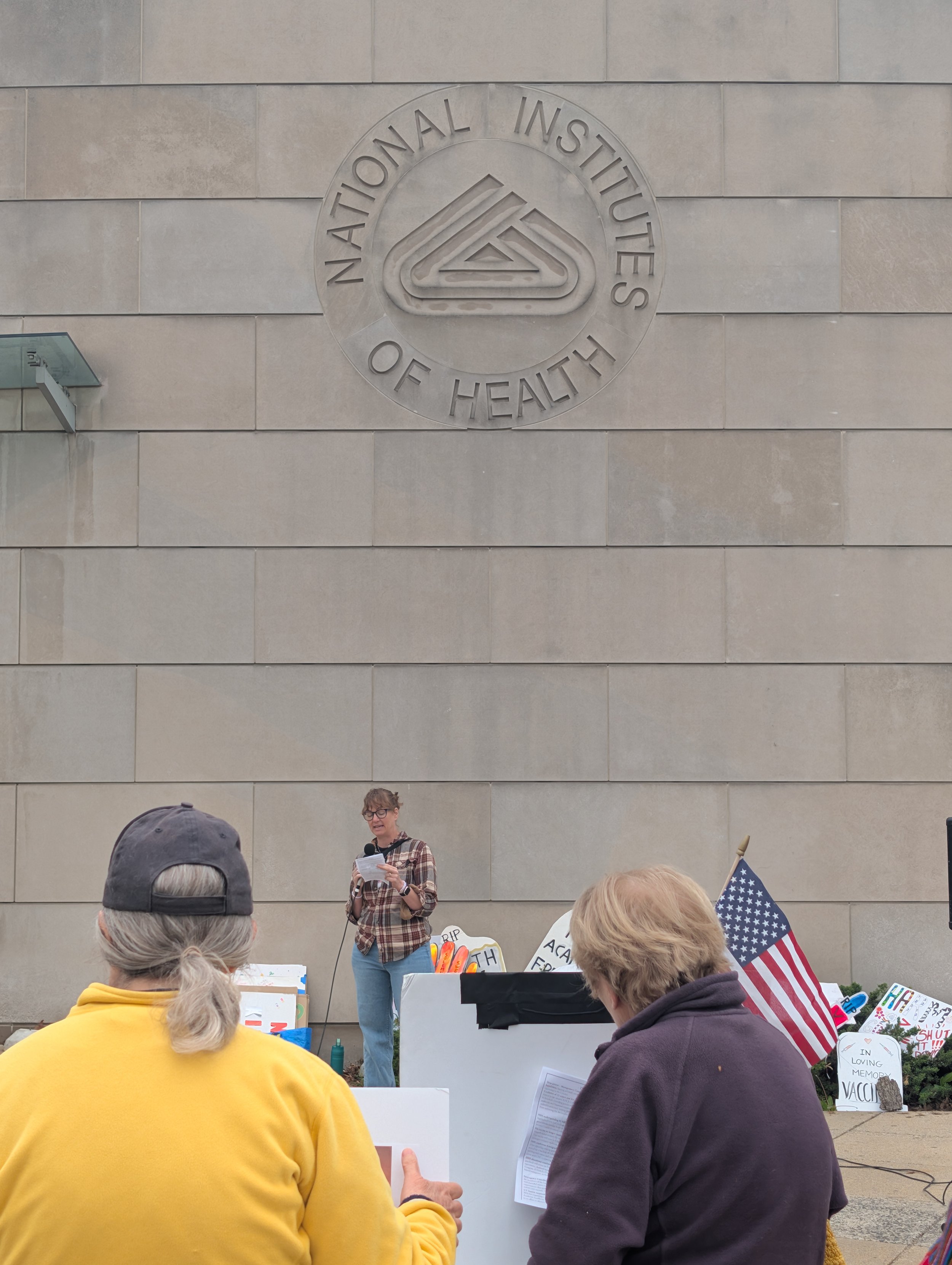 Vigil attendees hold an American flag watching the speaker in front of a building with the NIH logo