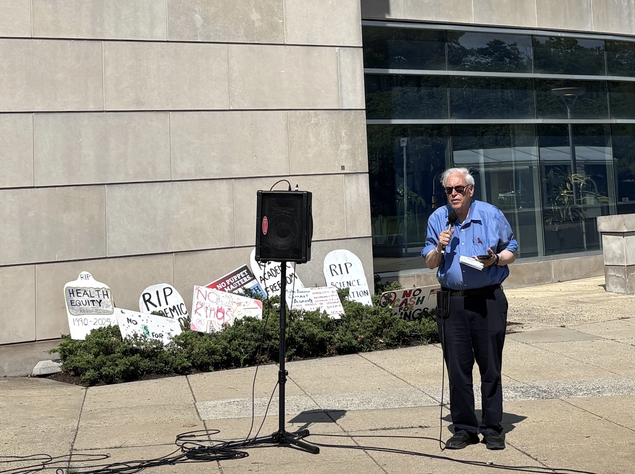 A man speaks into a microphone at the NIH Vigil beside signs styled as tombstones.