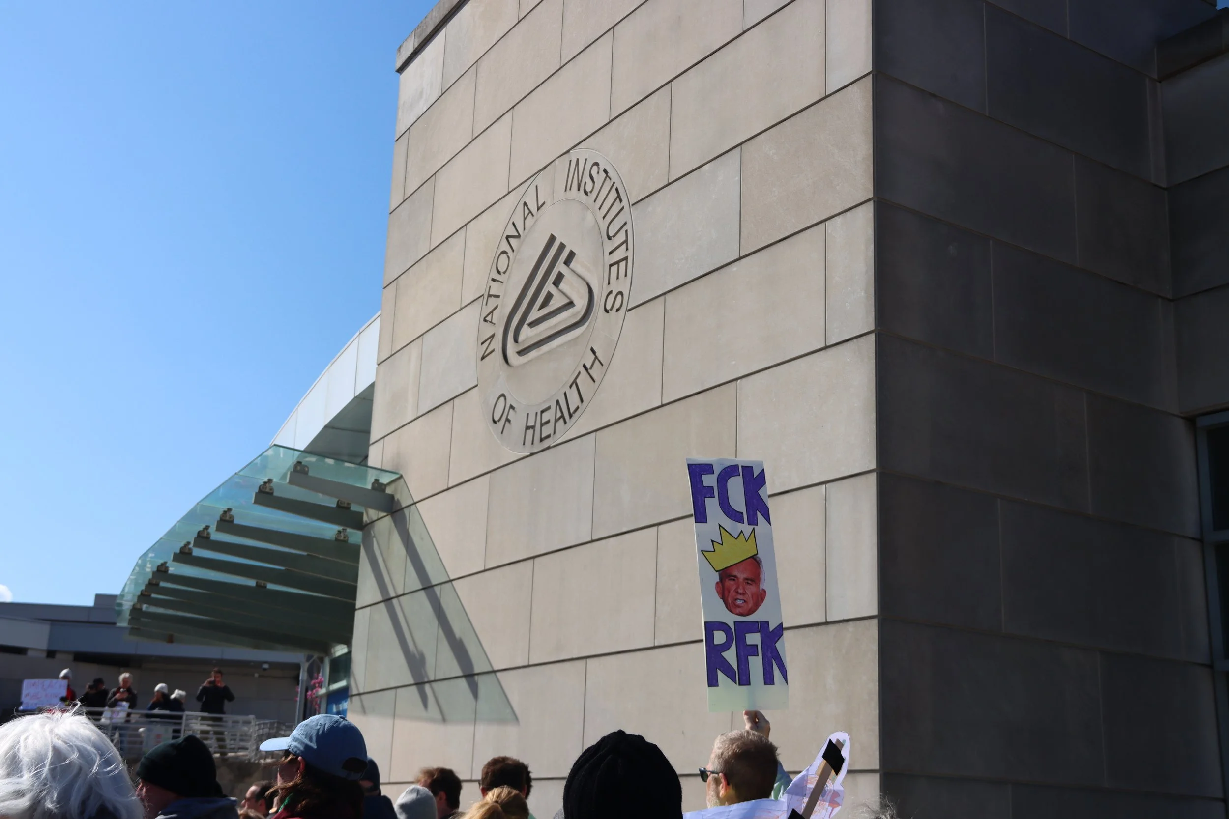 A "fck RFK" protest signs with the NIH logo on a building in the background