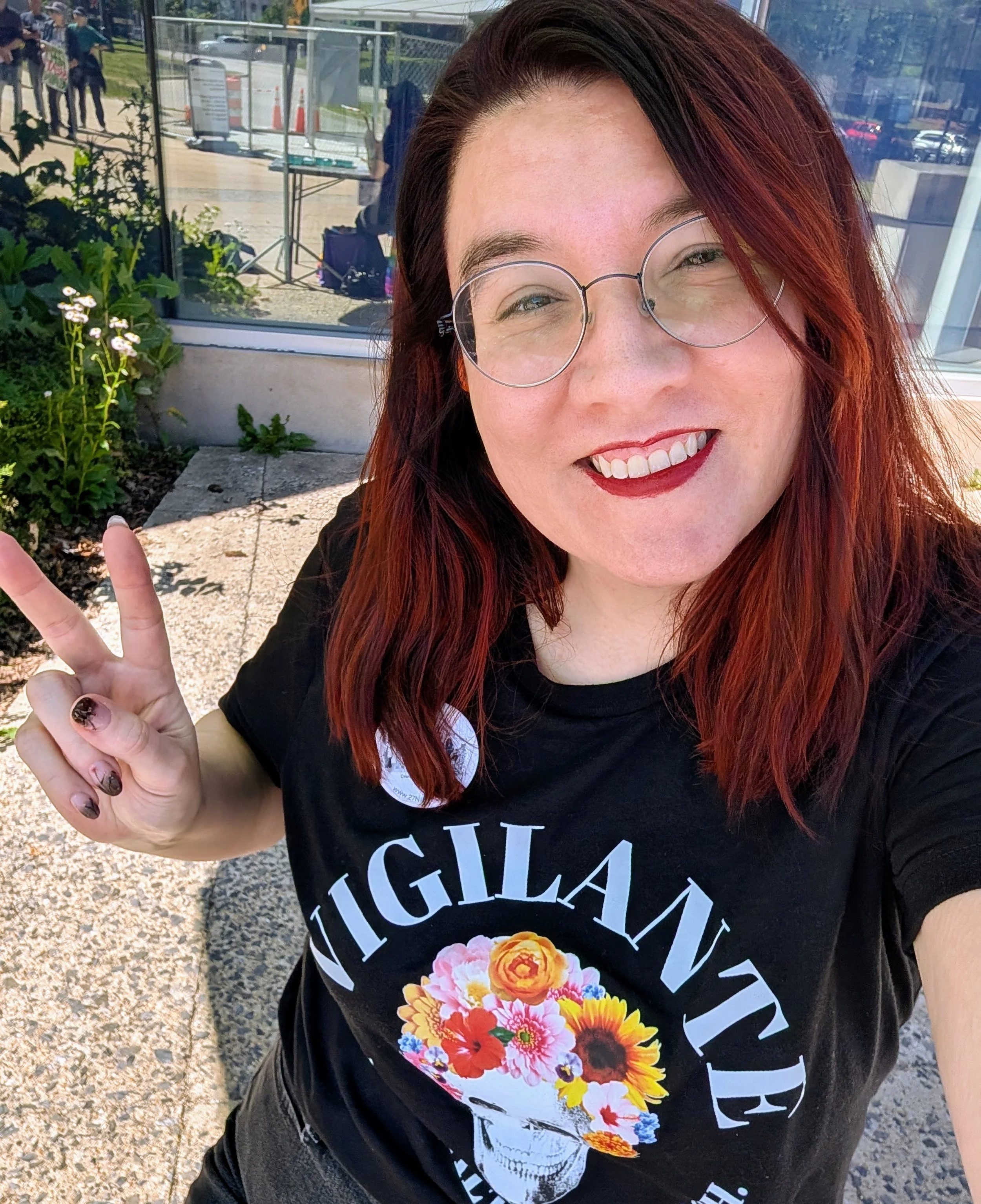 A woman in a vigil-themed Vigilante t-shirt smiles and flashes a peace sign