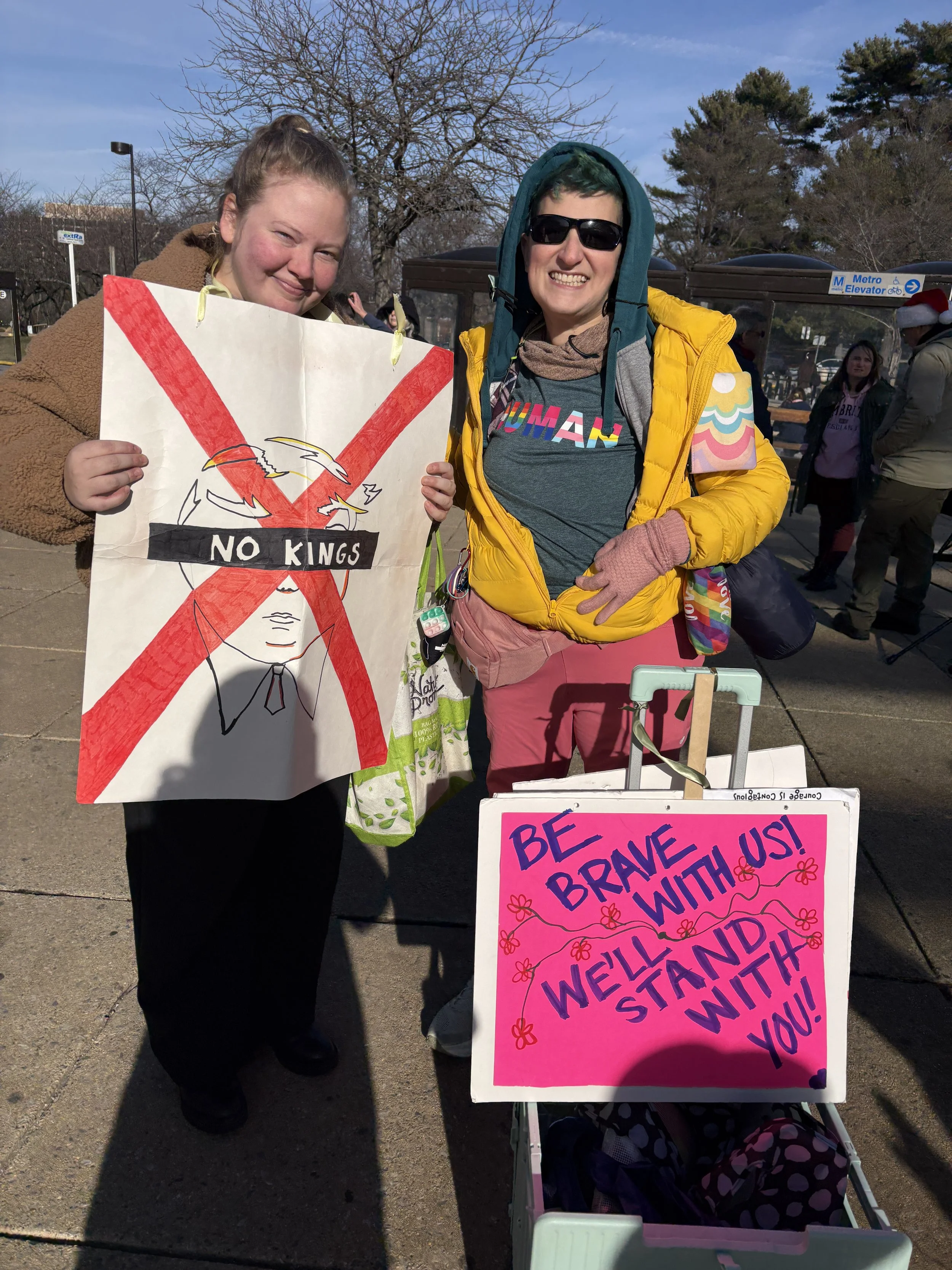 Two people holding protests signs reading "No Kings" and "Be Brave With Us, We'll Stand With You."
