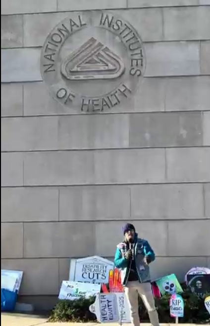 A person speaking at an NIH vigil, with the NIH logo visible on the wall above thema