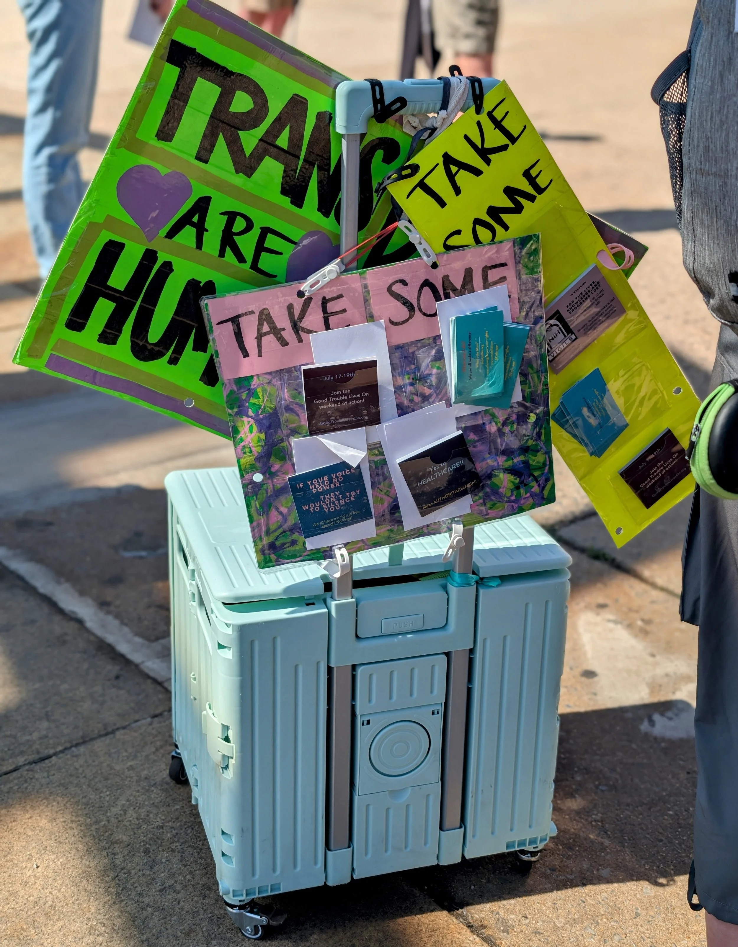 A rolling crate with protest signs and a card holder saying "Take some"