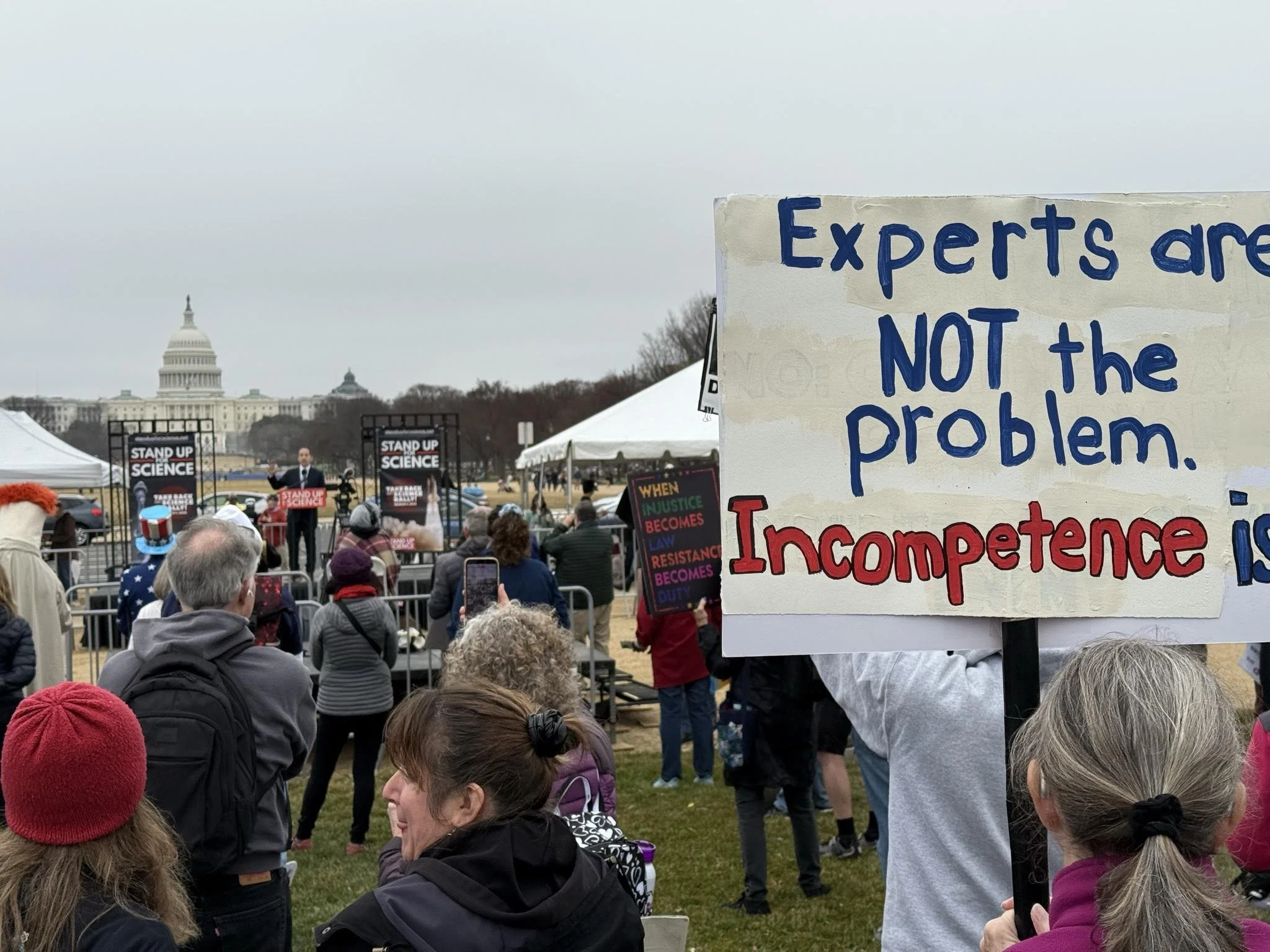 The rally crowd, including a protest signs that says "Experts are not the problem. Incompetence is."