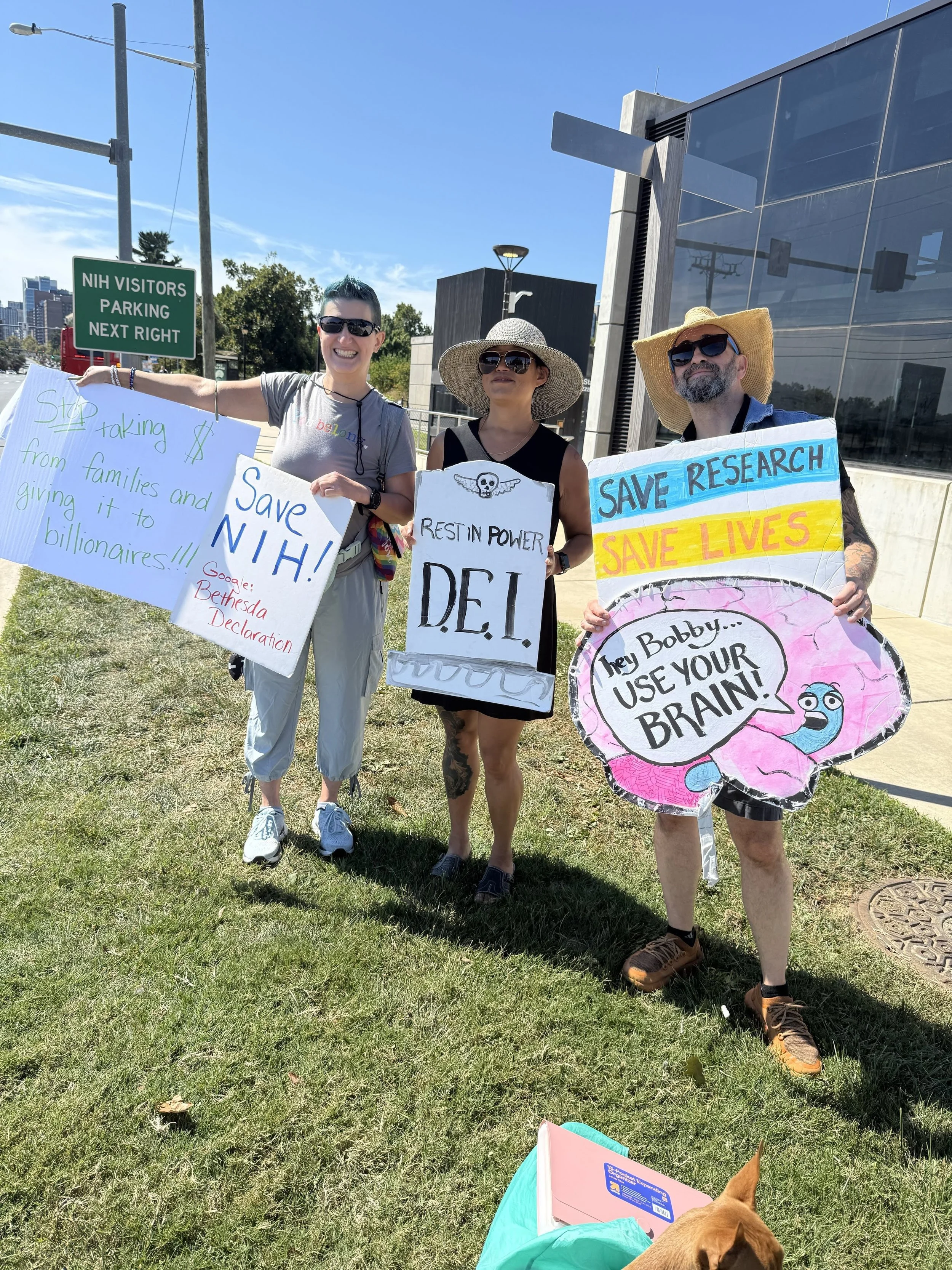 A group of three people holding pro-science protest signs while standing on Rockville Pike near the NIH campus.