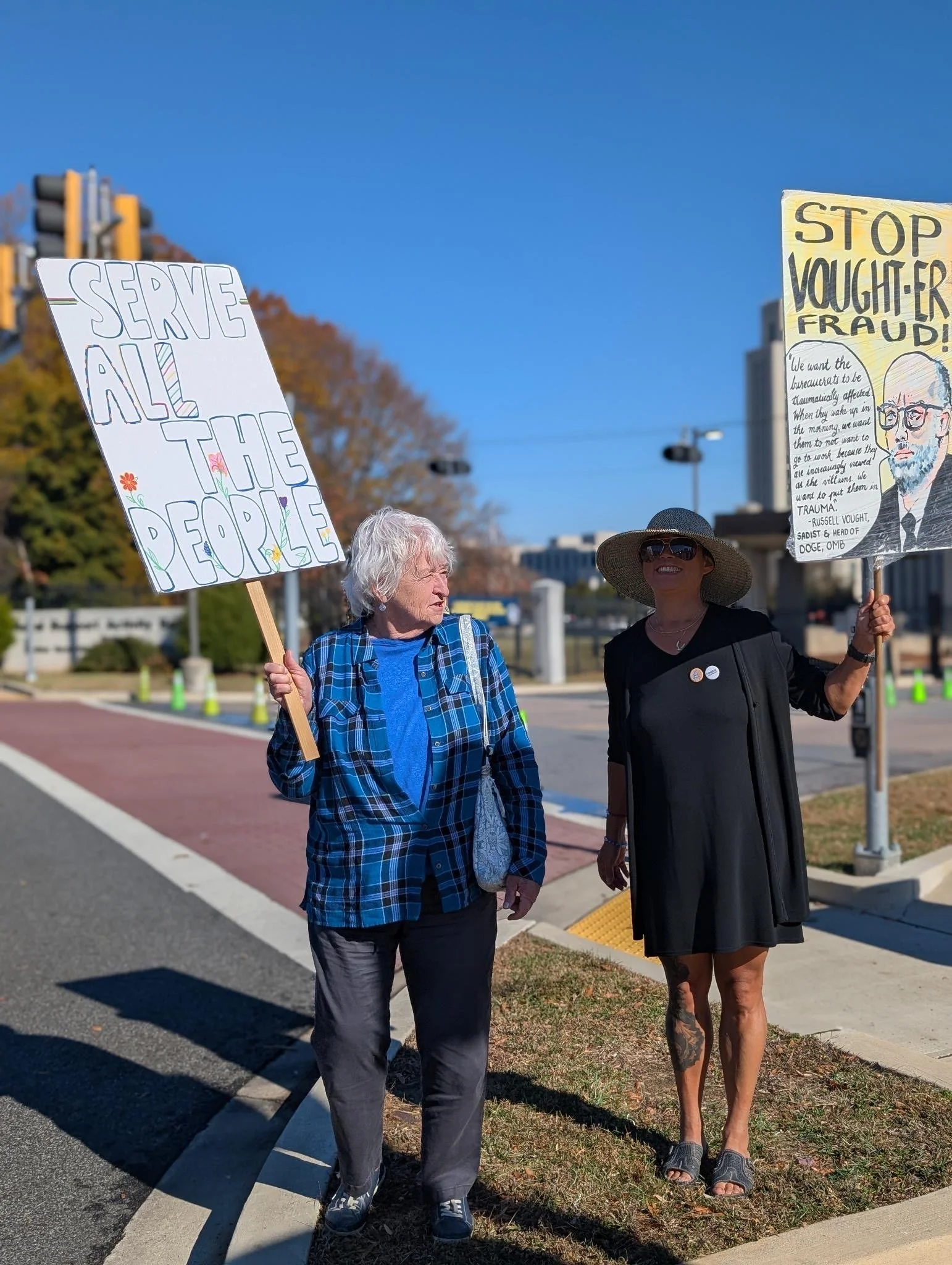 Two women stand by a road, holding protest signs reading "Serve all the people" and "Stop Vought-er fraud."