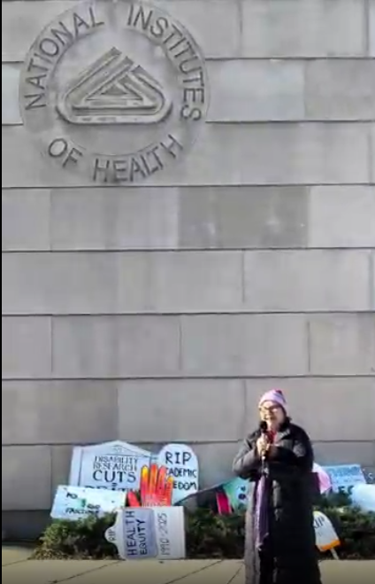 A person speaking at an NIH vigil, with the NIH logo visible on the wall above thema
