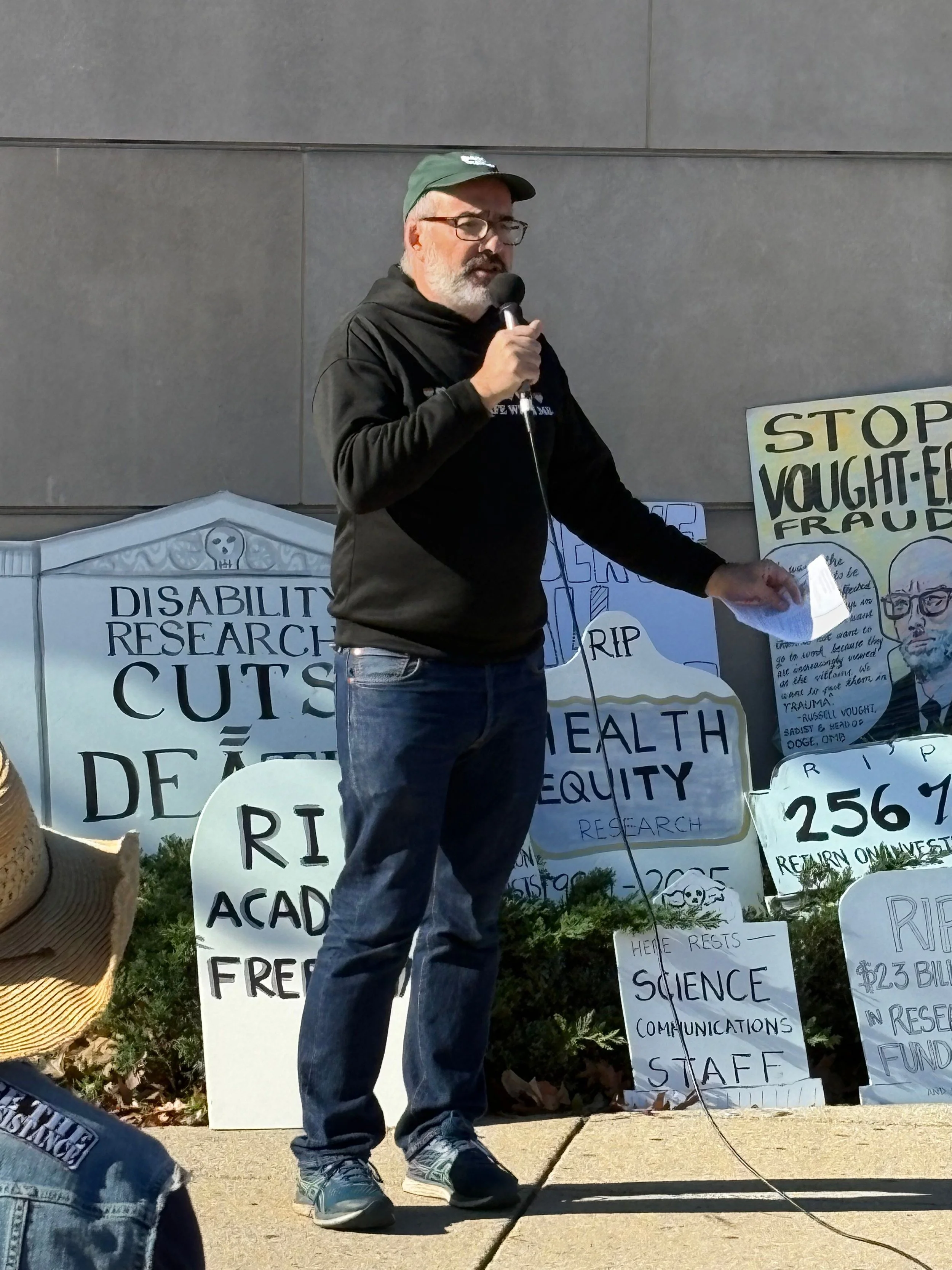 A man speaks into a microphone, standing in front of a line of protest signs