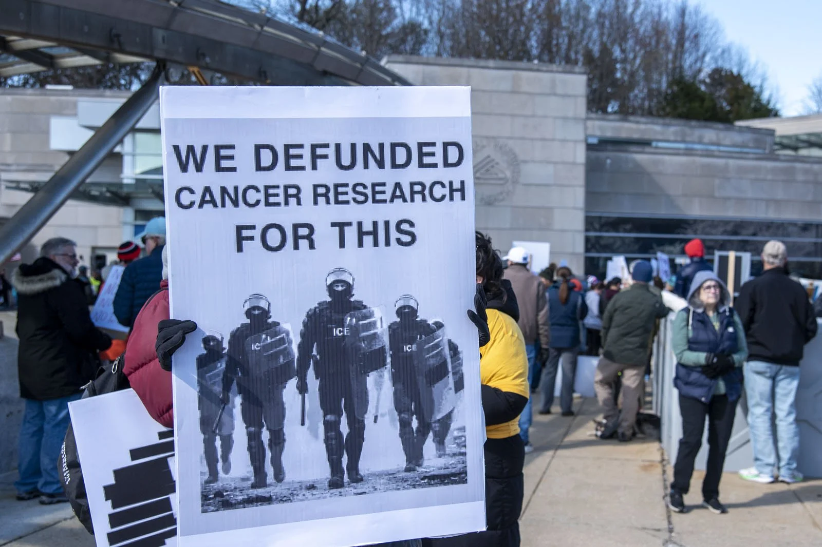 A person holding a protest sign showing ICE agents with the caption "We defunded cancer research for this"