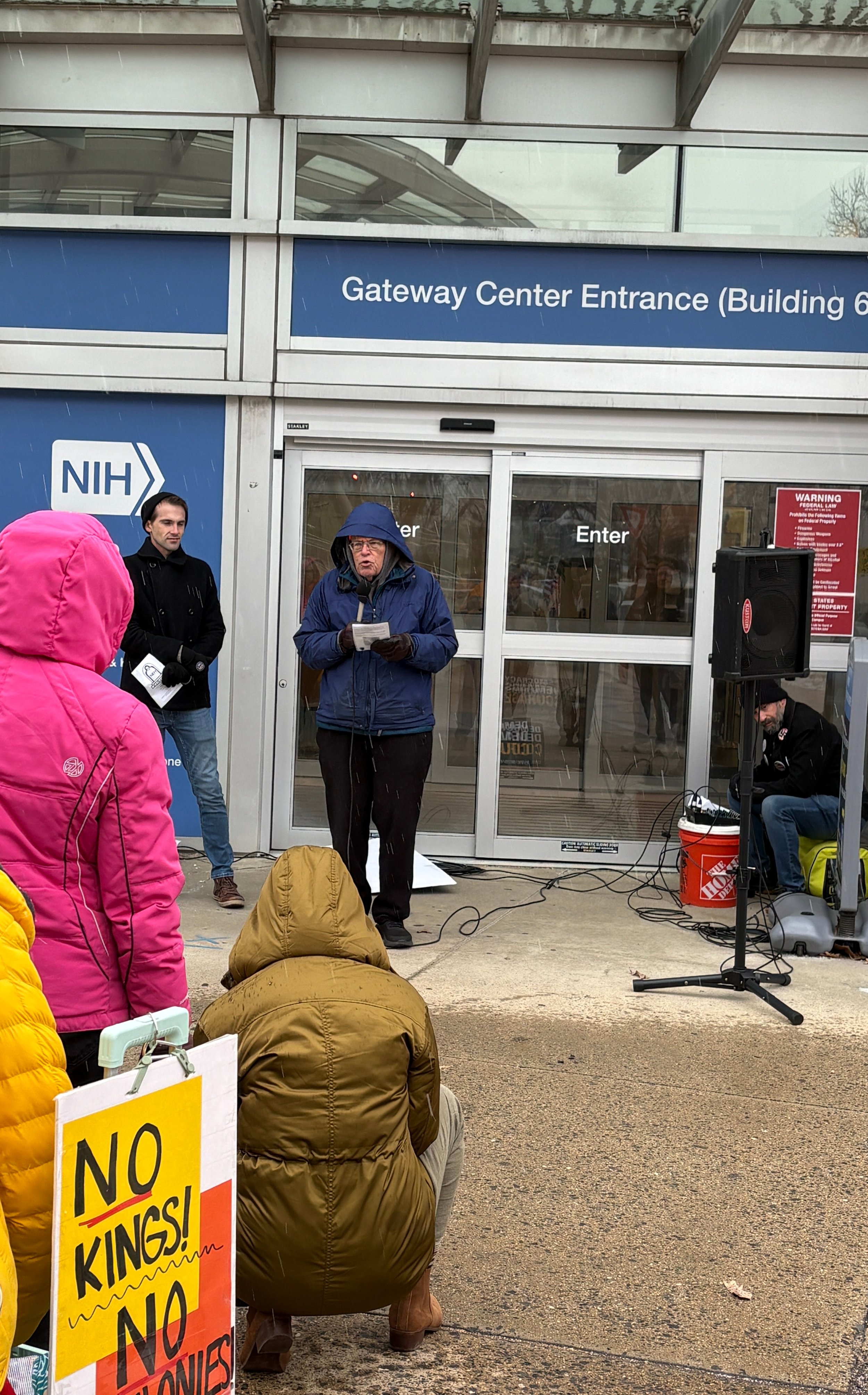 A person speaks at an NIH vigil, with a "No Kings! No Colonies!" protest sign in the foreground