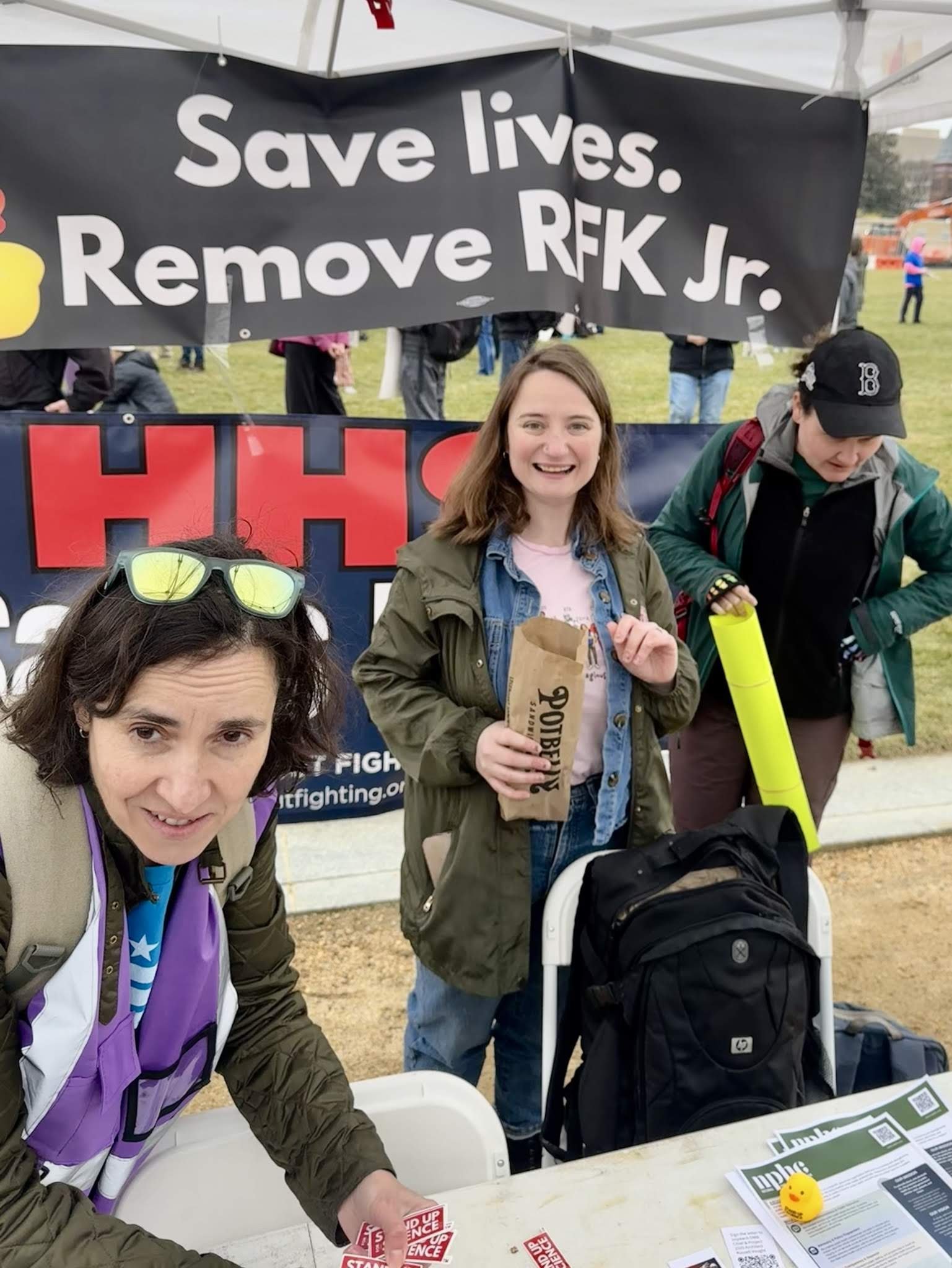 Three people at a table with a "Save lives. Remove RFK Jr." sign.