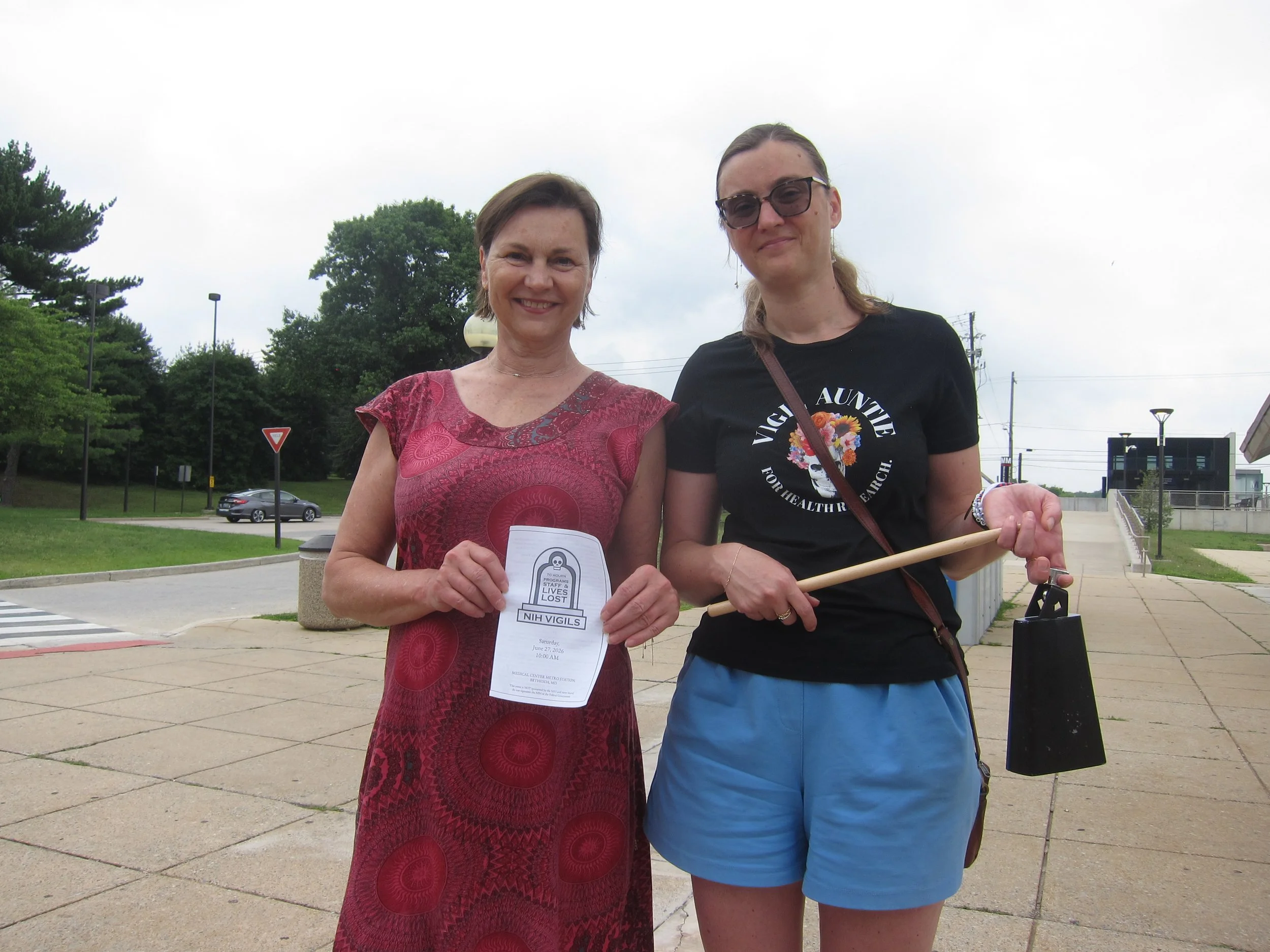 A woman holding a vigil program and another woman wearing a Vigil Auntie shirt and holding a cowbell
