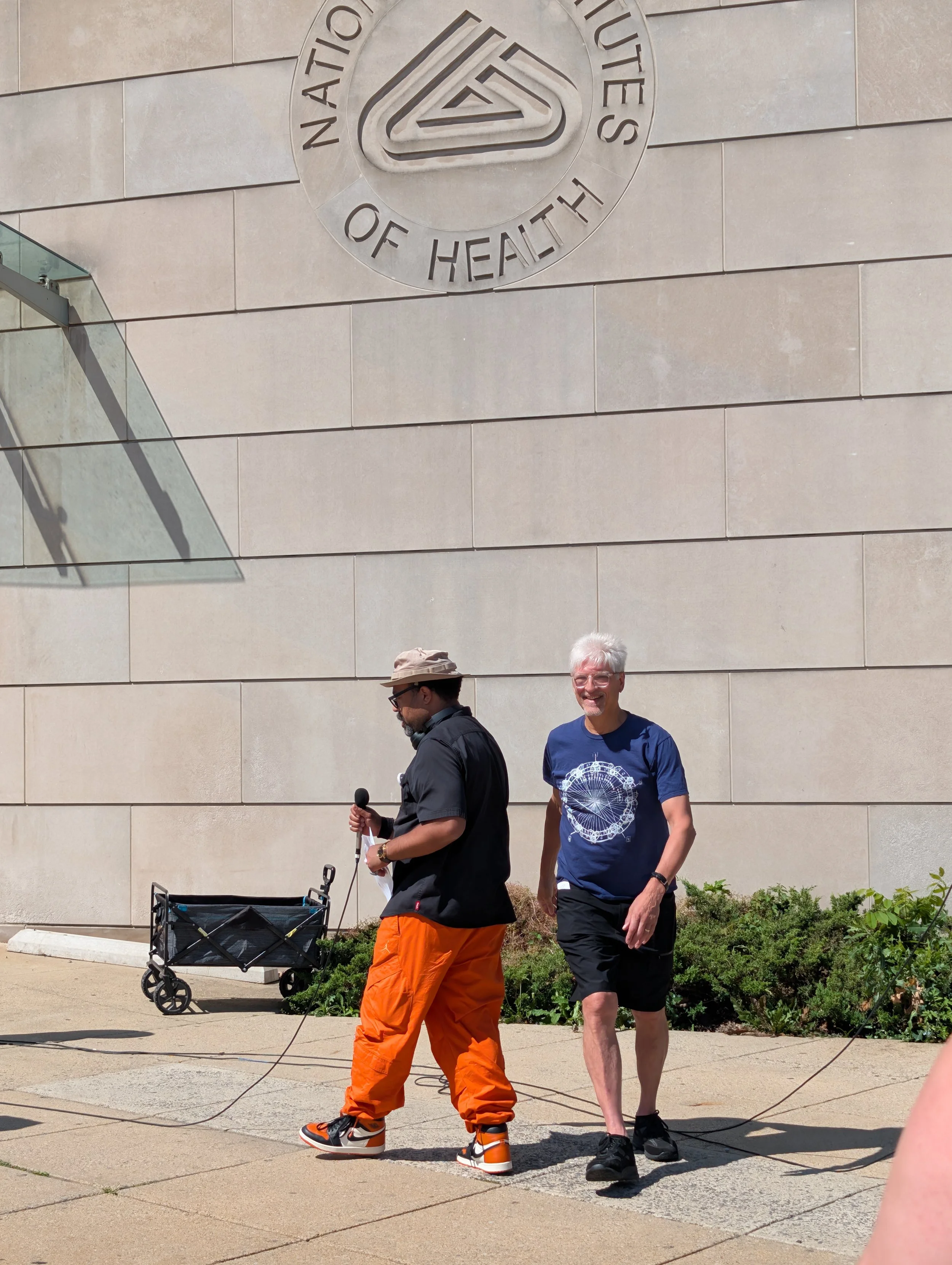 Two NIH Vigil speakers stand near a microphone beneath the National Institutes of Health seal.