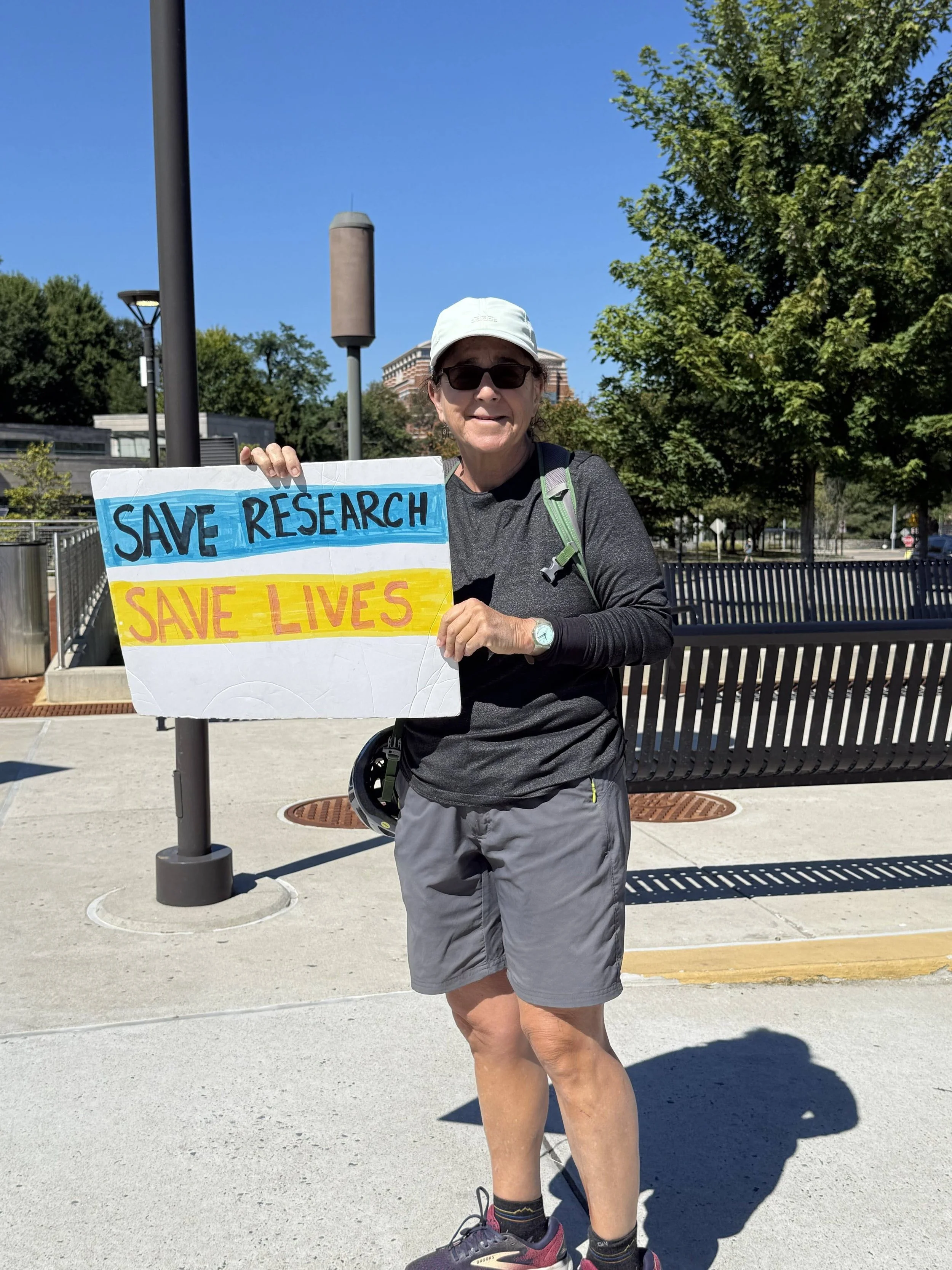 A person in a white baseball cap and dark gray clothing holding a protest sign that says Save Research, Save Lives.